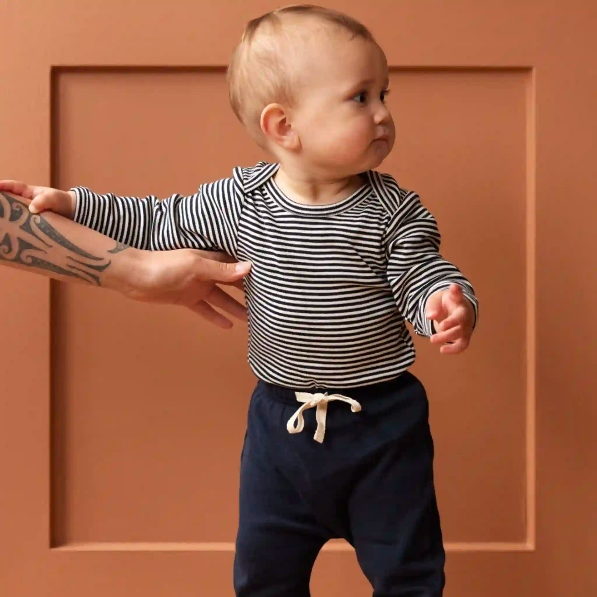 Baby in a striped shirt and navy pants standing against a brown wall.