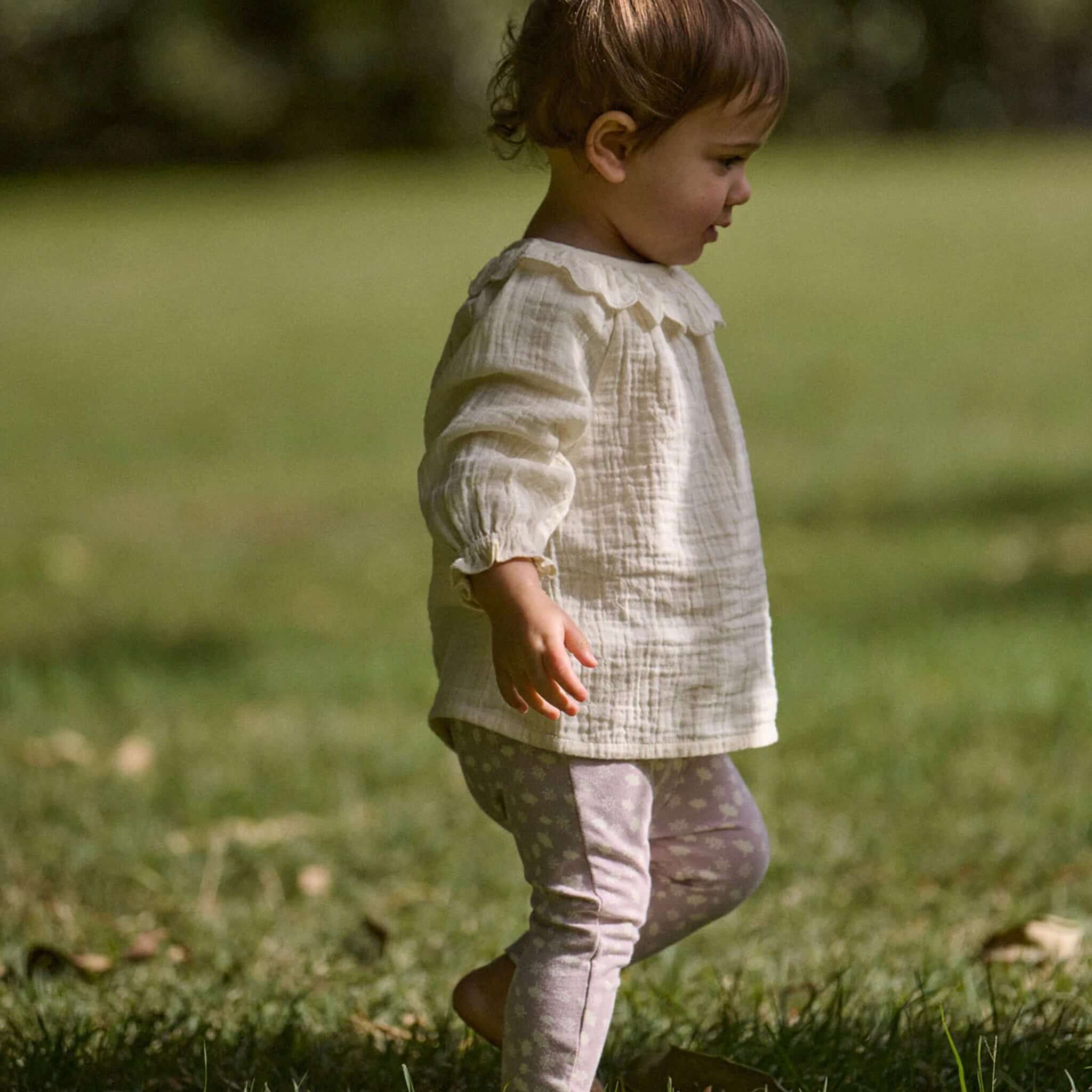 Nature Baby Organic Cotton Ruffle Cameron Blouse - Natural worn by a toddler playing in a grassy field.
