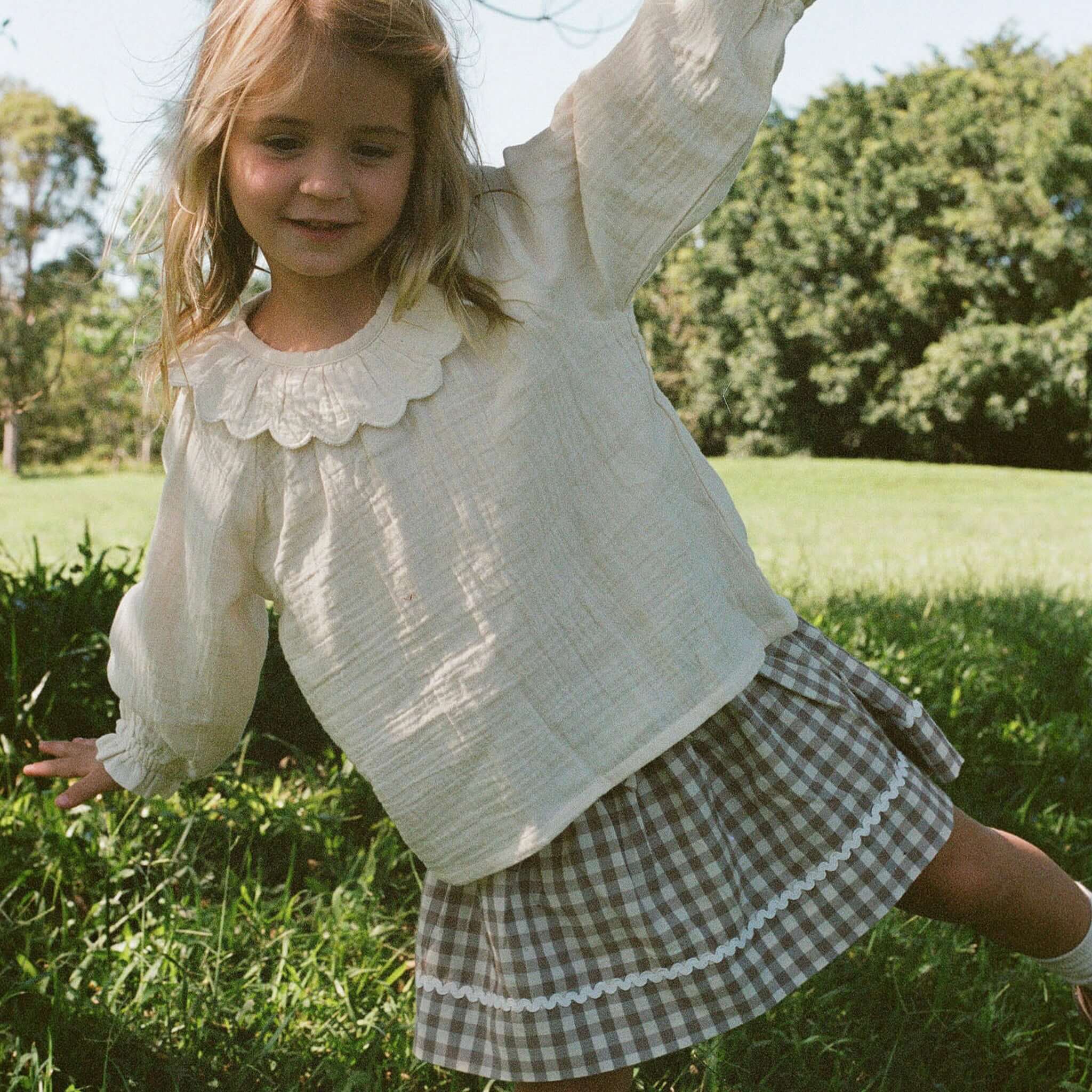 Nature Baby Organic Cotton Ruffle Cameron Blouse - Natural worn by a girl playing outdoors in a grass field.