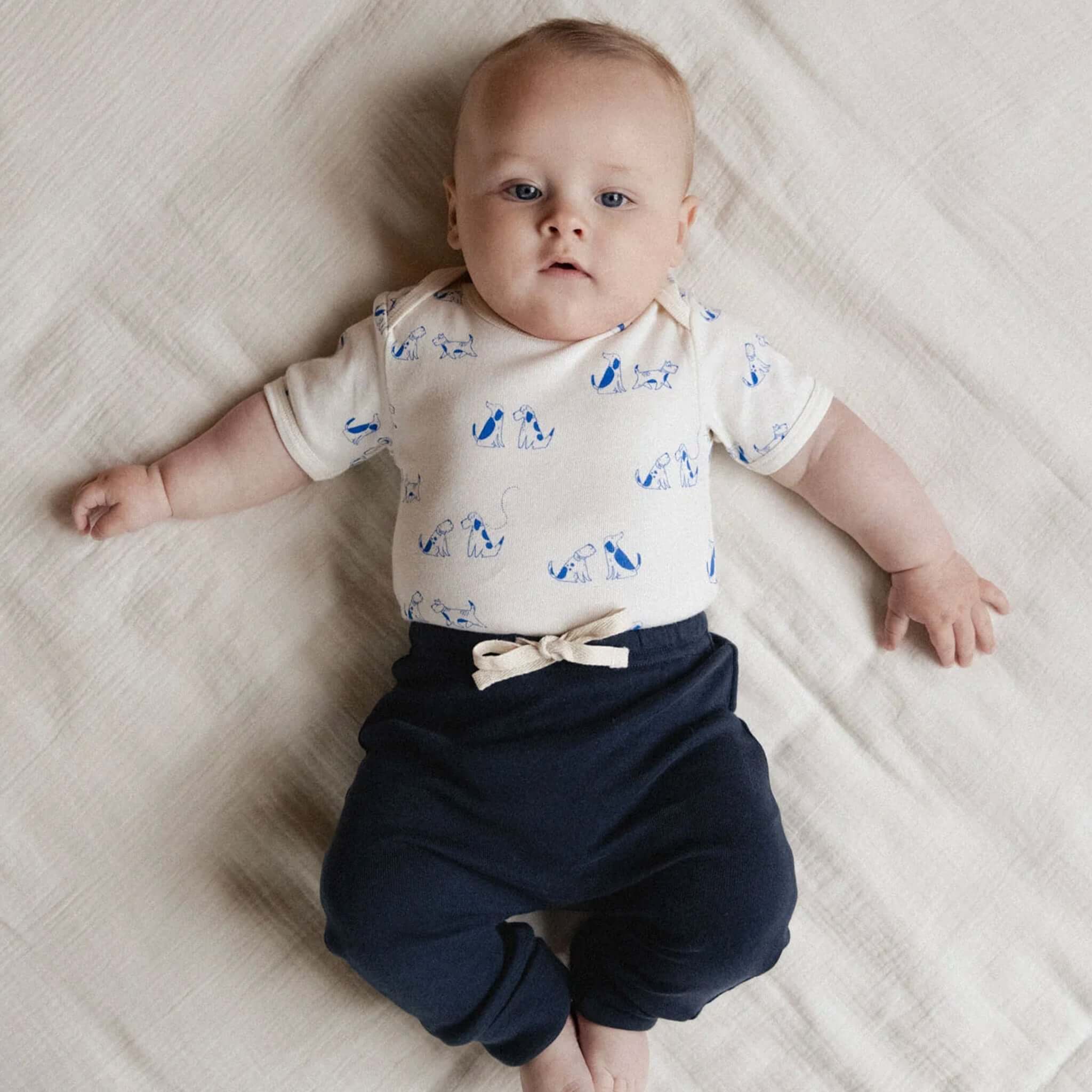 Nature Baby Organic Cotton Short Sleeve Bodysuit - Dog Walk worn by a baby lying on a blanket, showcasing dog prints.