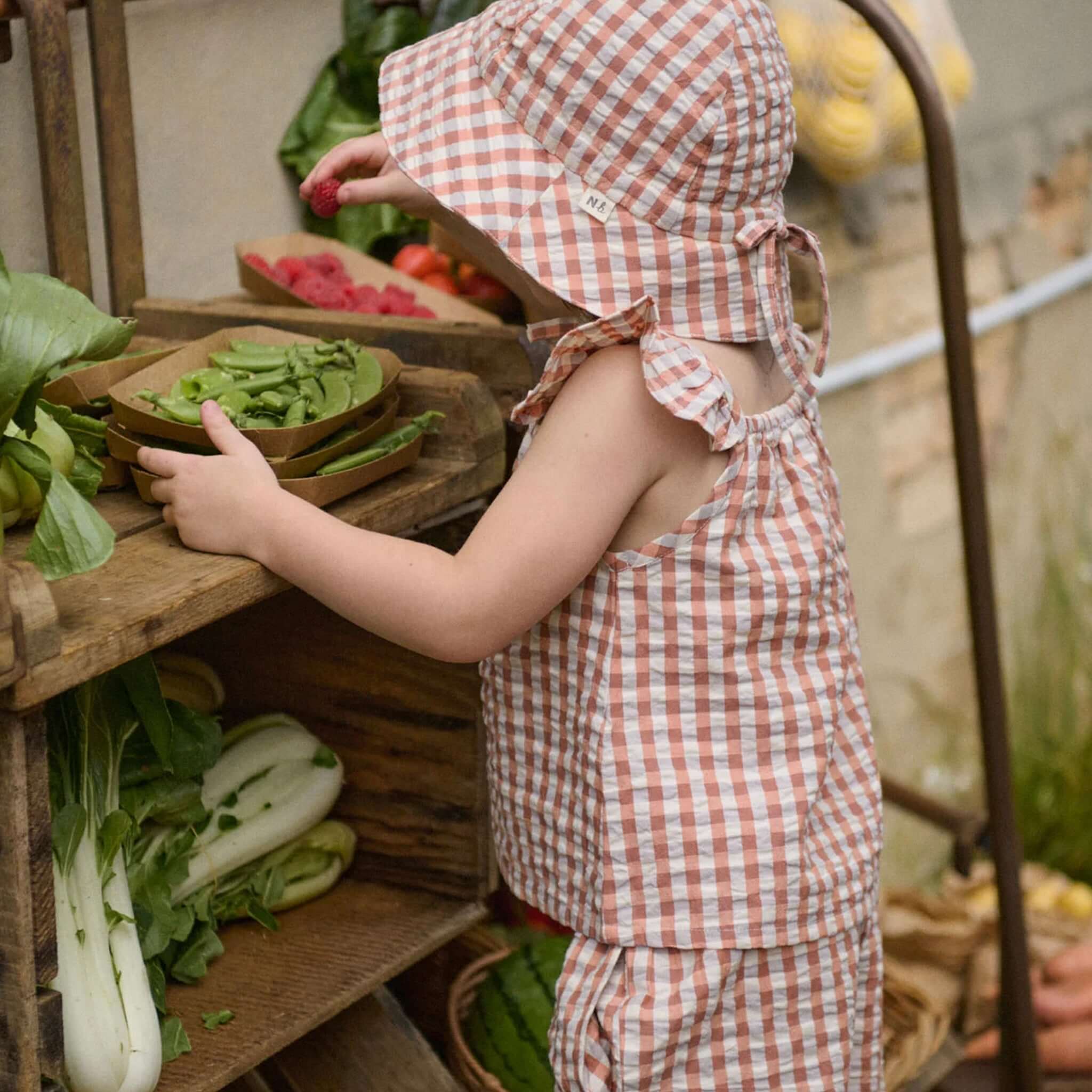 Child wearing Nature Baby Organic Cotton Sunhat - Dandelion Gingham, exploring fresh vegetables on a wooden shelf.