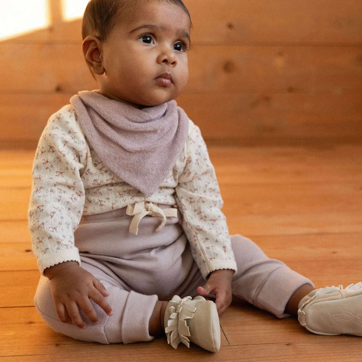 Nature Baby Organic Cotton Terry Triangle Bib - Lilac worn by a baby sitting on wooden floor, styled with soft pastel outfit.