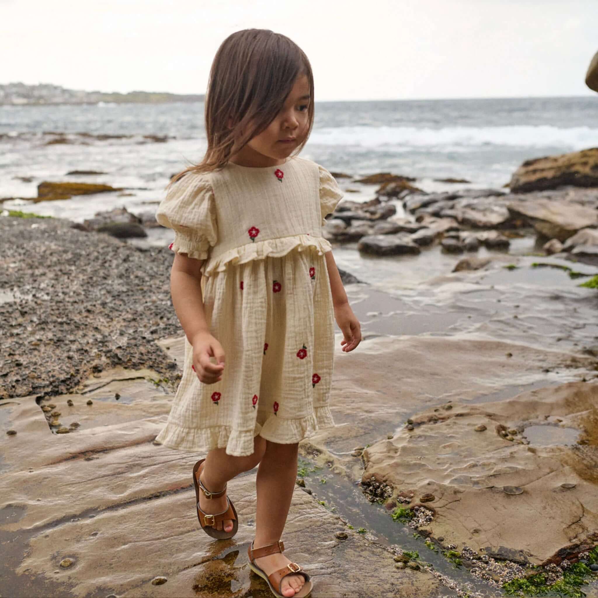 Nature Baby Organic Cotton Valentina Dress - Red Flora, girl walking on rocks by the ocean in floral dress.