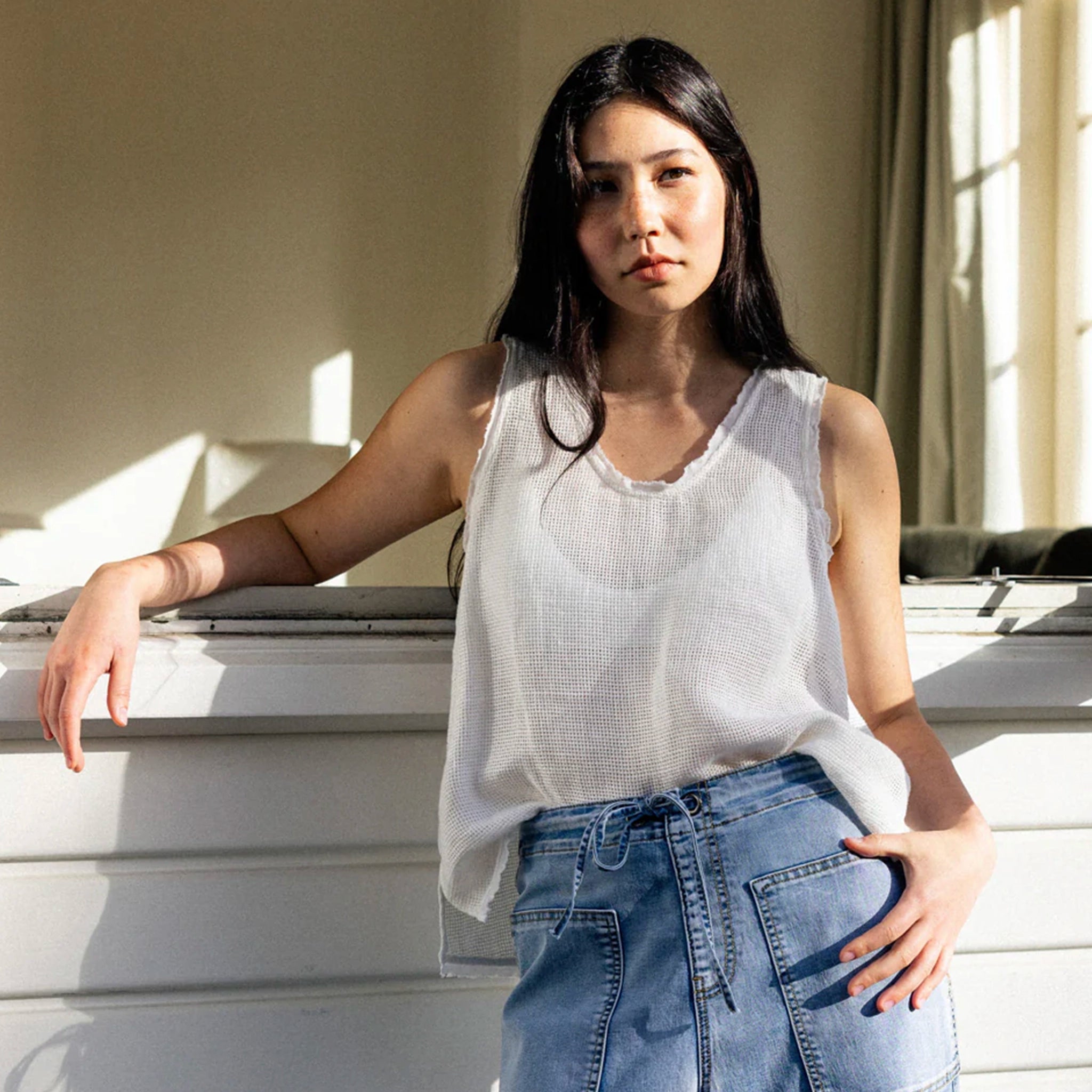 Woman wearing a white sleeveless top and blue jeans standing indoors.