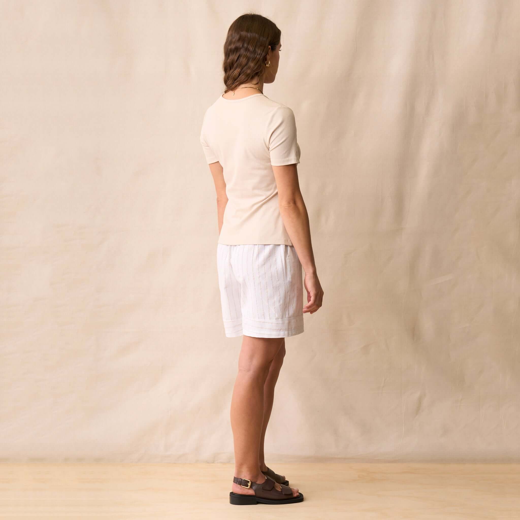 A woman wearing the Neris Ziggy Tee - Sand with linen shorts, viewed from the back.