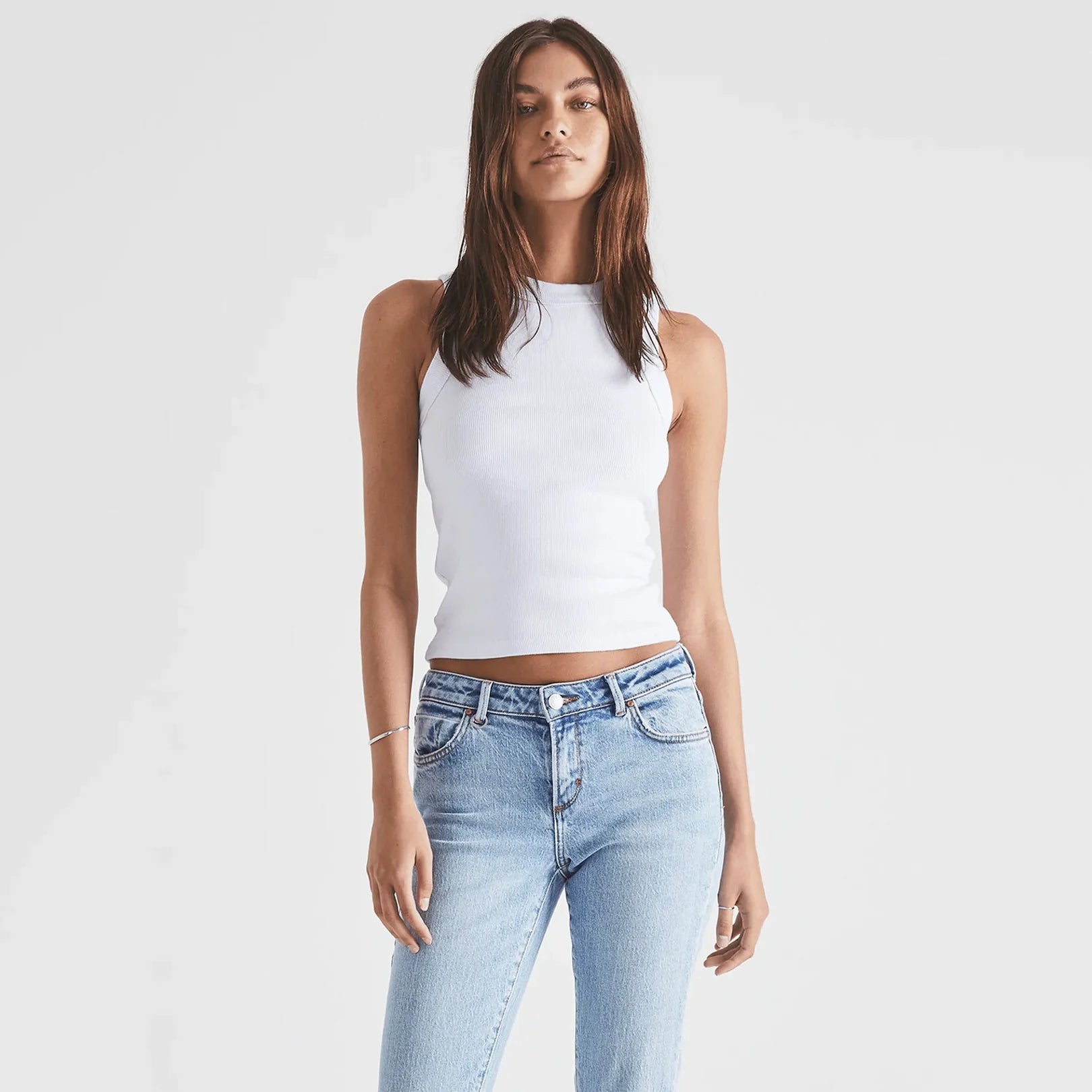 Woman wearing a white sleeveless top and light blue jeans on a plain background