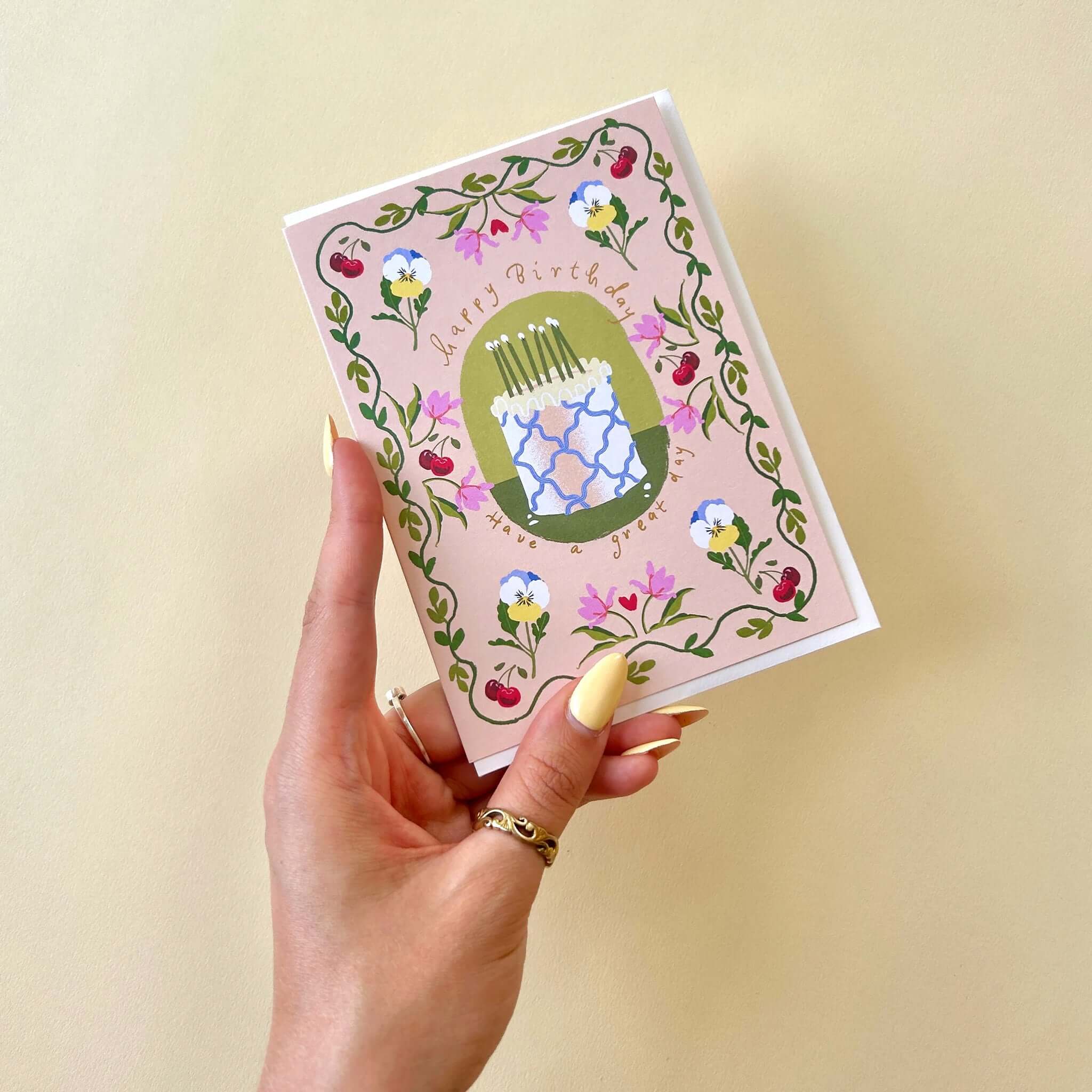 Noi Publishing Card - Cake and Cherries Birthday held by a hand, featuring an iced cake and floral border.