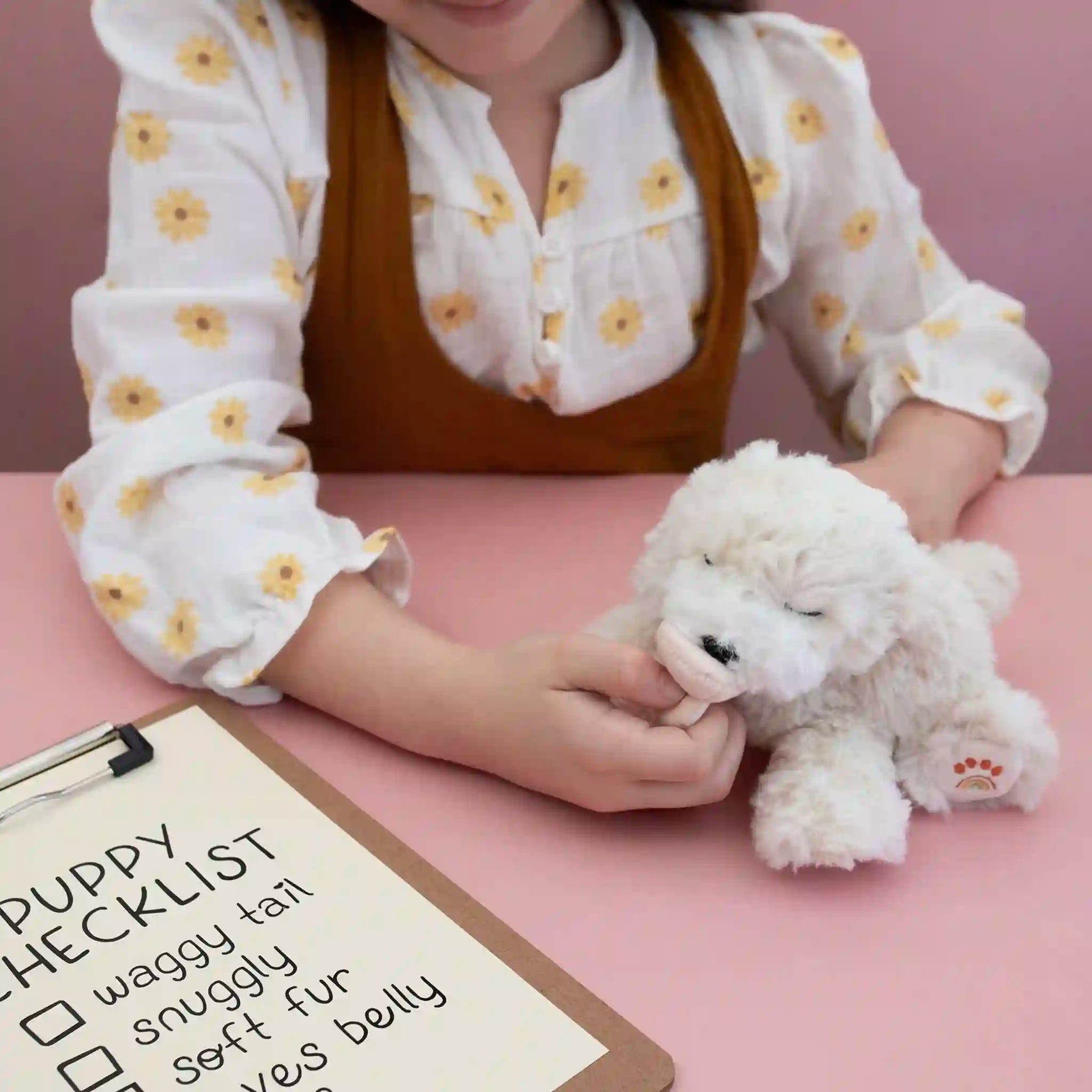 Person holding a small white dog plush toy with a checklist on a pink surface