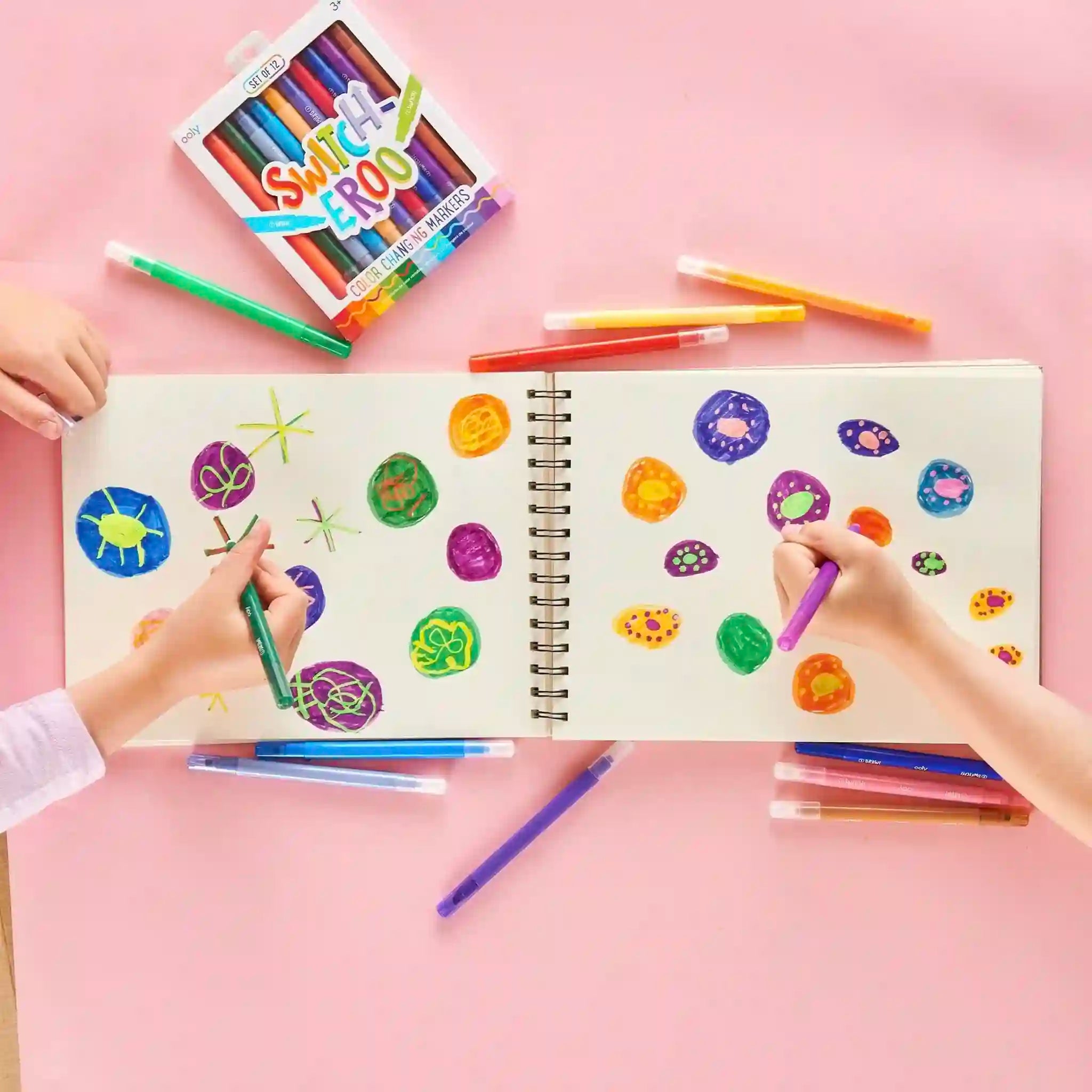 Children coloring in a spiral-bound notebook with colorful markers on a pink background