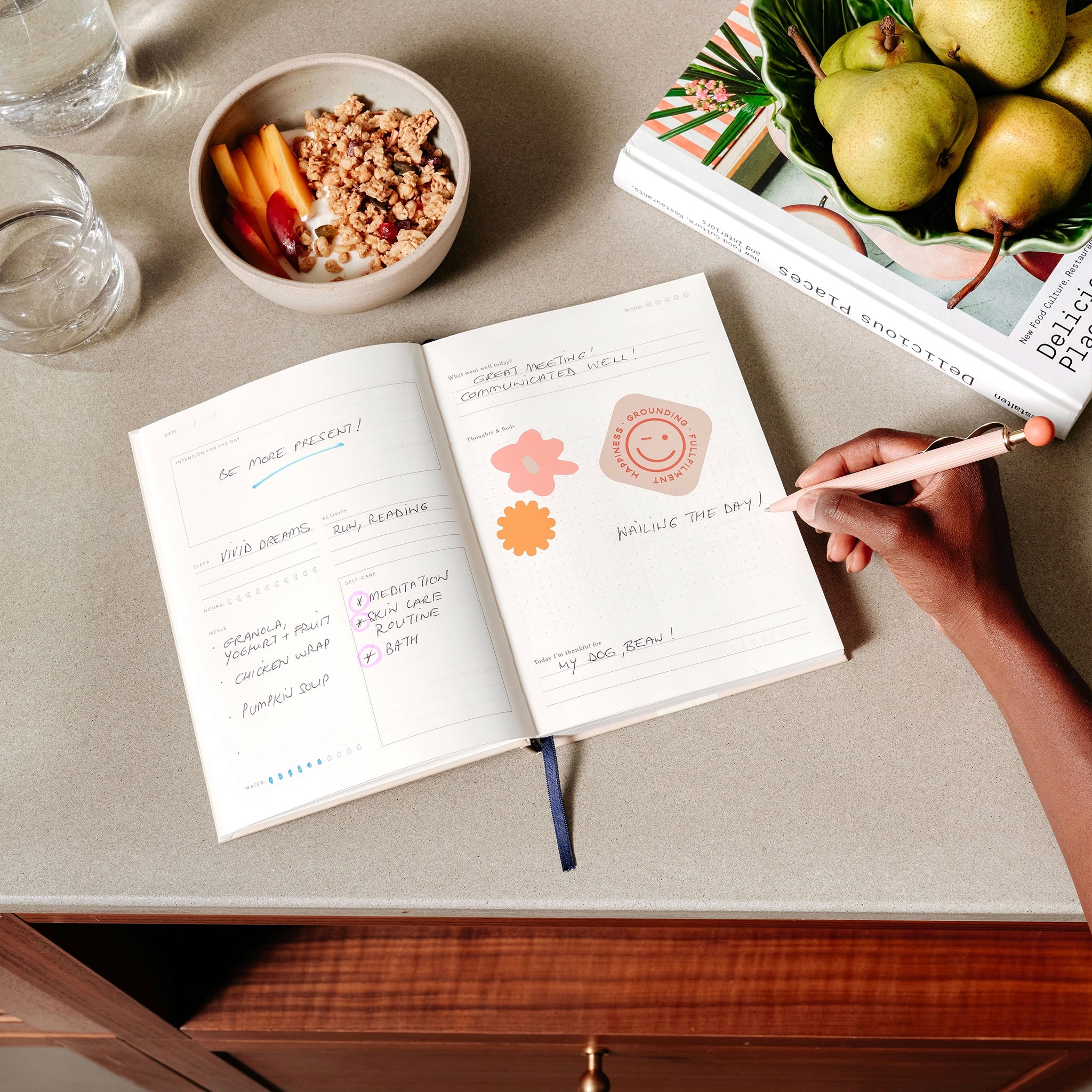 Person writing in a notebook on a table with a bowl of fruit and a book in the background
