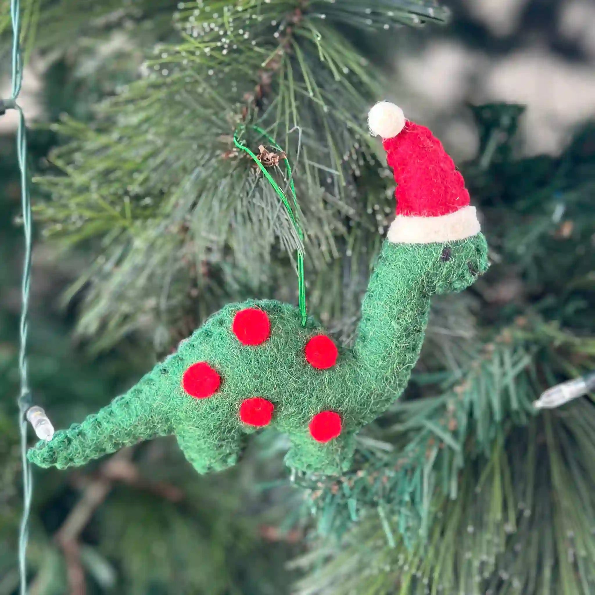 Green dinosaur ornament with red polka dots and a Santa hat hanging on a Christmas tree.