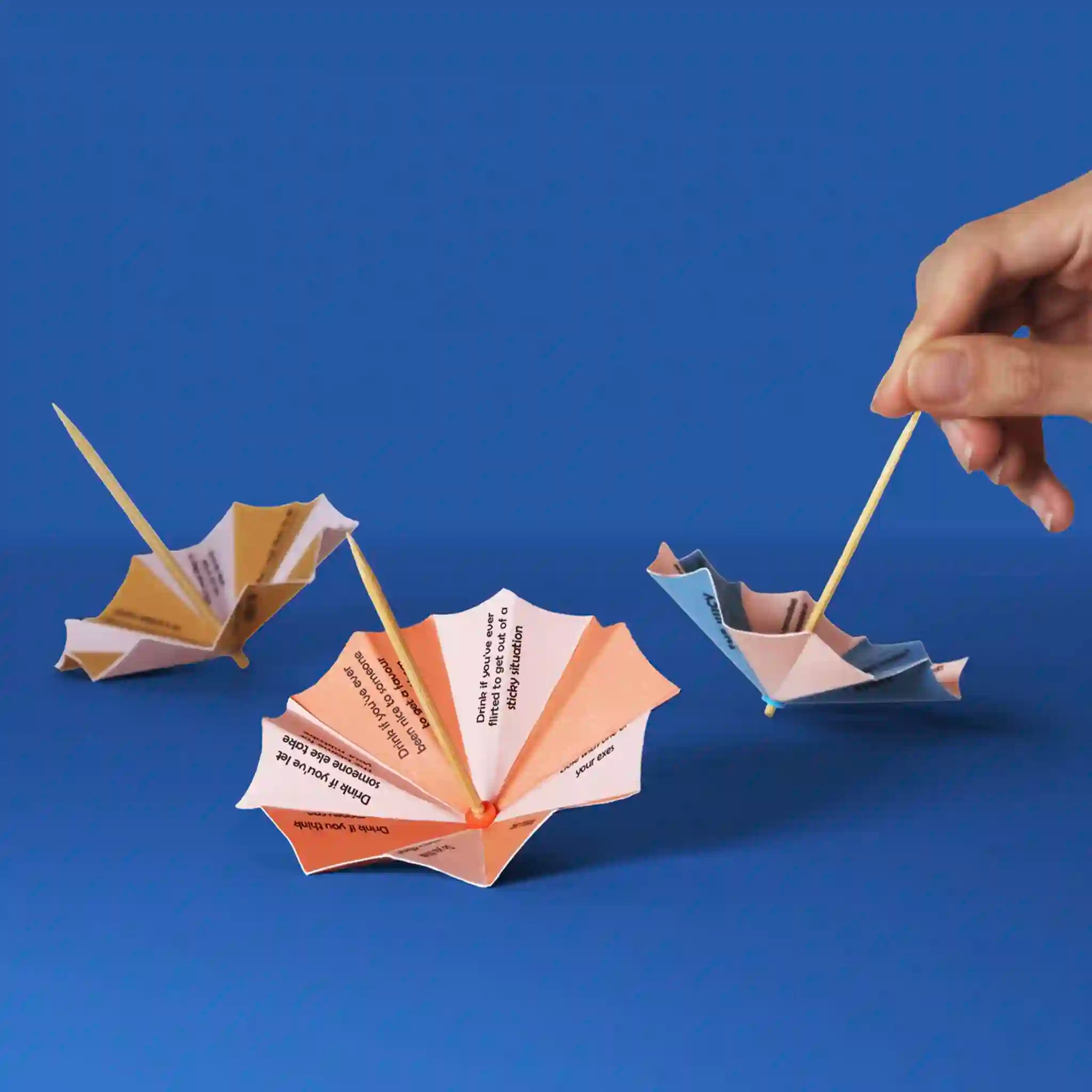 Three paper umbrellas with toothpicks on a blue background, one being adjusted by a hand.