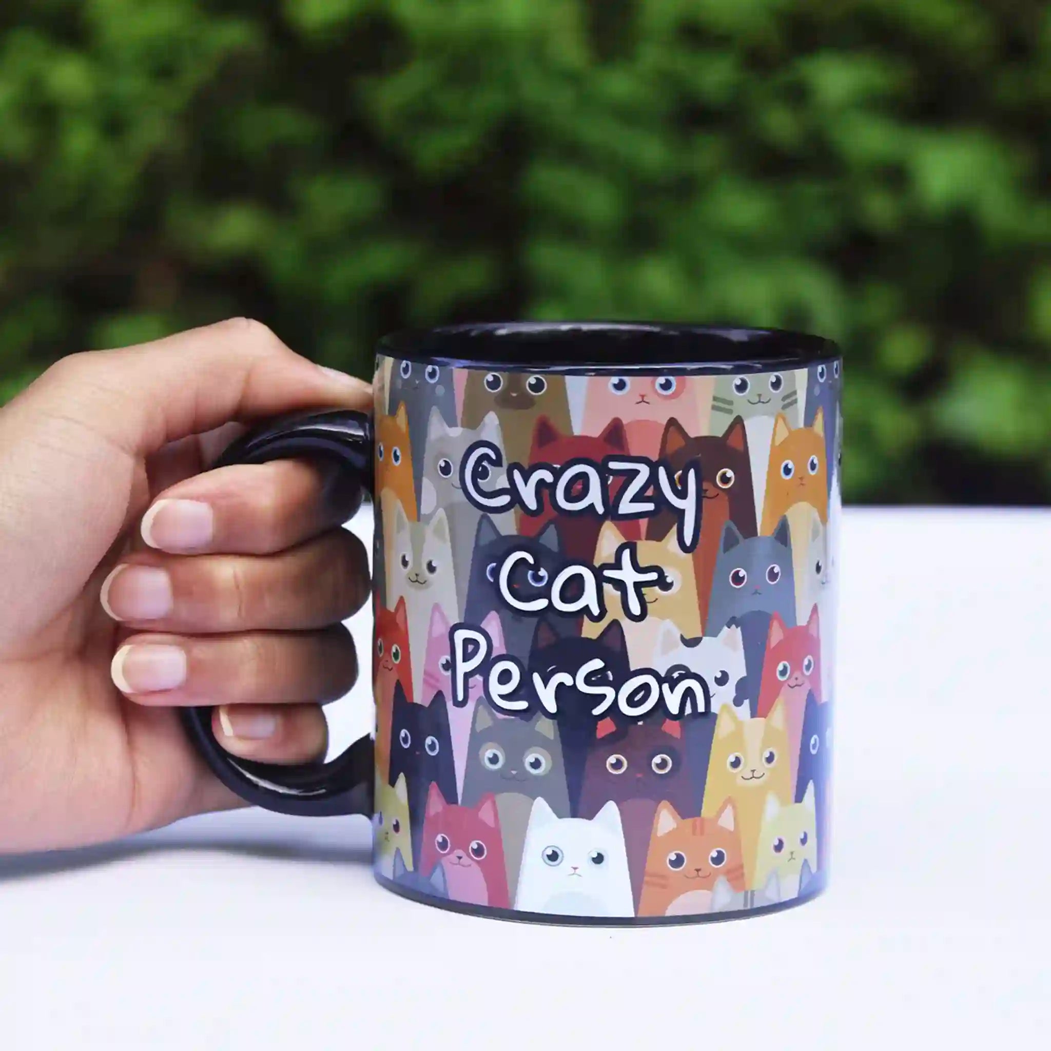 Mug with cat pattern and 'Crazy Cat Person' text held by a hand outdoors.