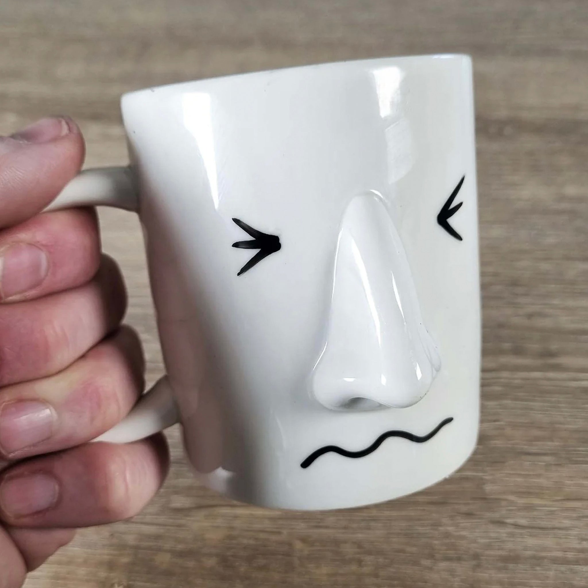 White mug with a face drawing held by a hand on a wooden surface