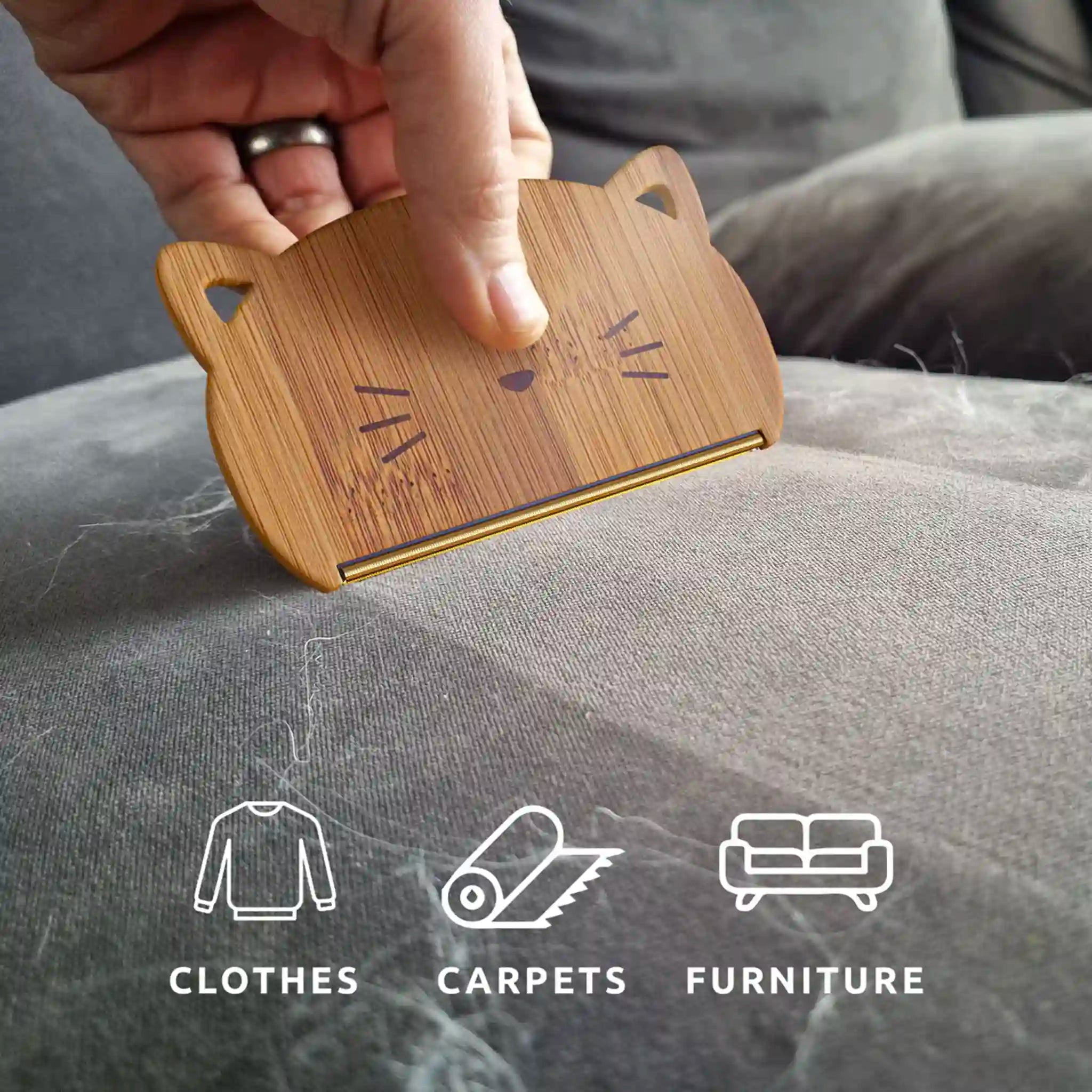 Wooden pet grooming brush with cat face design held over a textured surface, with icons below indicating use on clothes, carpets, and furniture.