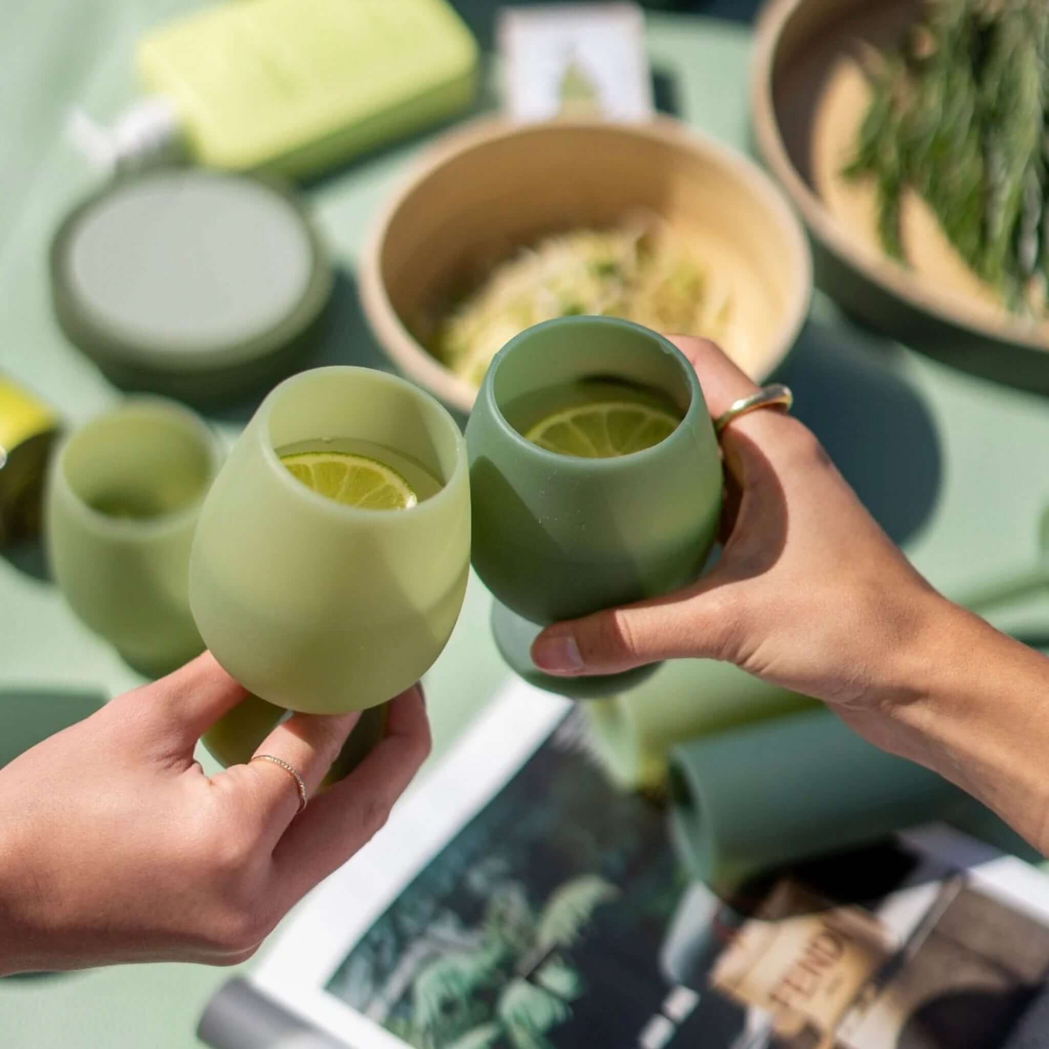 Two hands holding Porter Green Stemm Unbreakable Wine Glass Set - Sage & Olive with lemon slices inside.