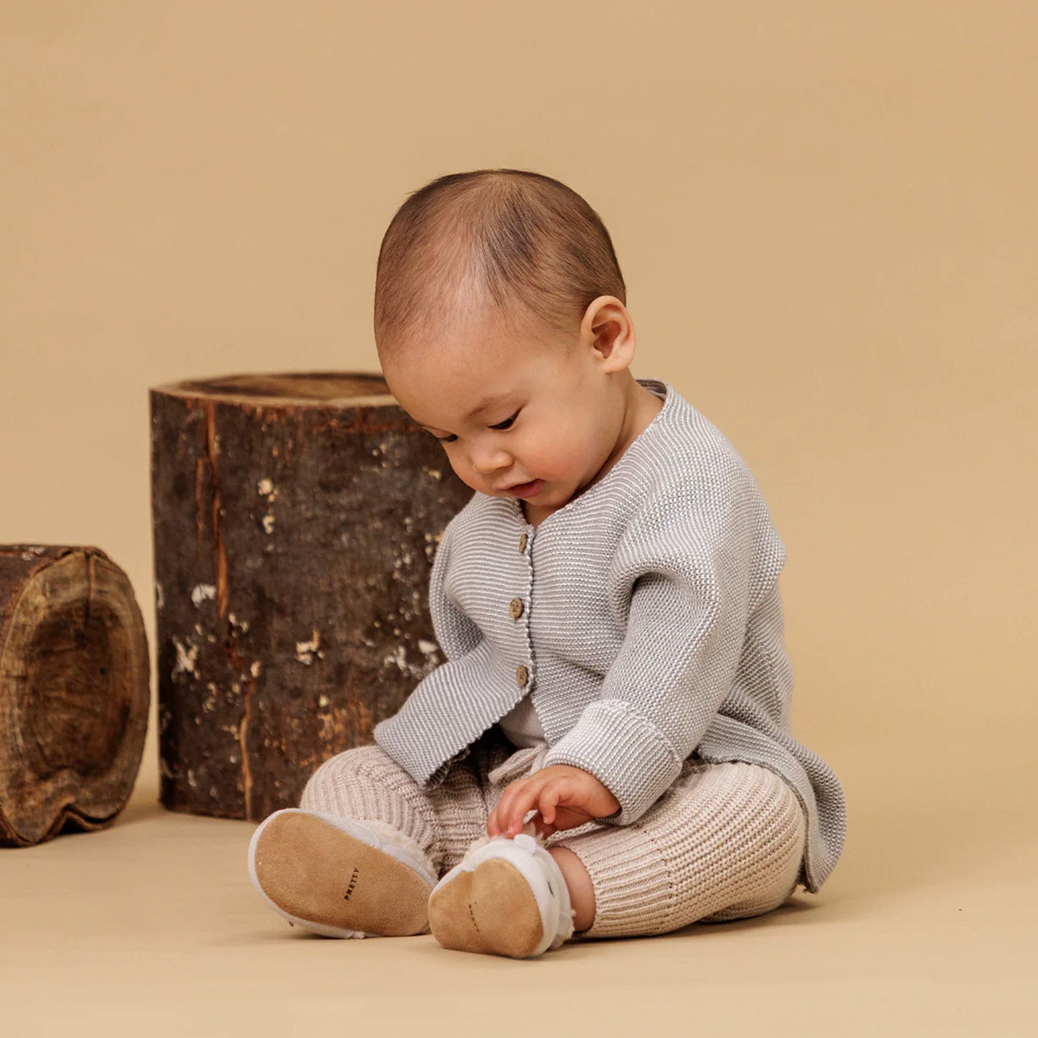 Baby in a light gray knitted outfit sitting on a beige surface with wooden logs in the background.