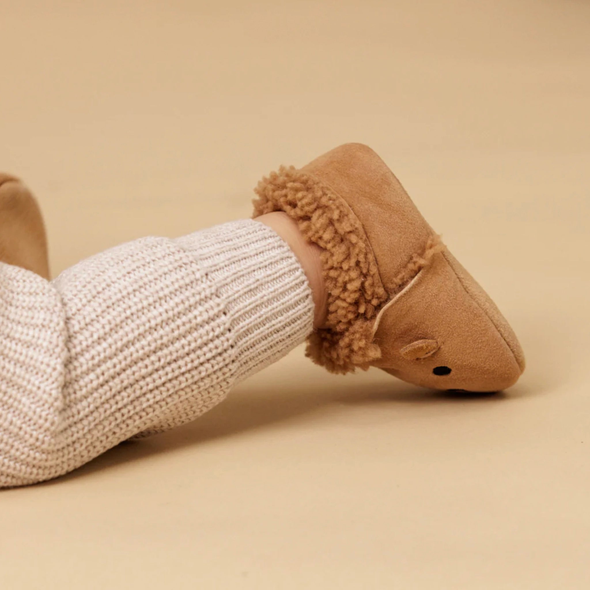 Tan slipper with fuzzy trim worn by a person in a white knit sock on a beige background
