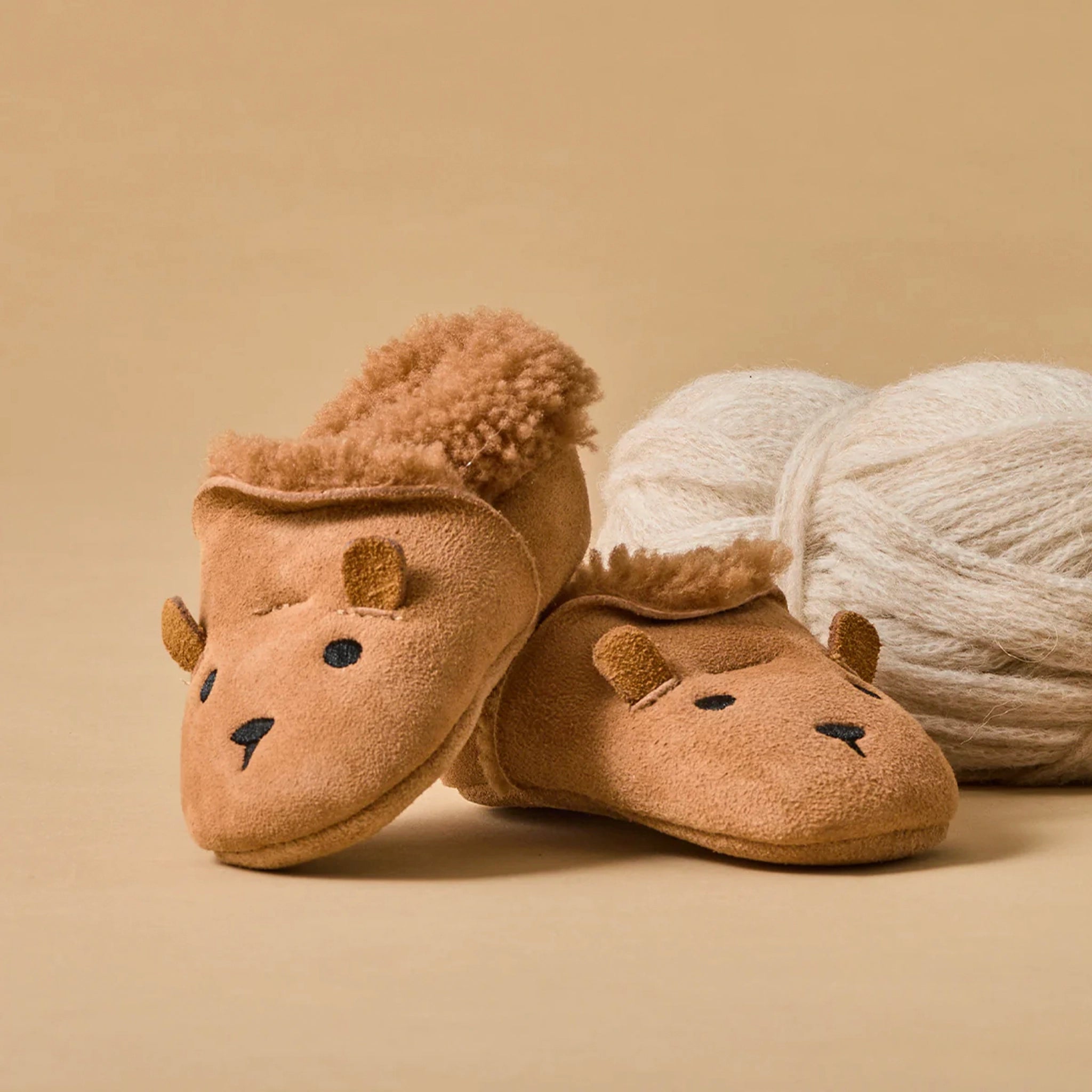Pair of brown bear slippers with fluffy texture next to a ball of yarn on a beige background