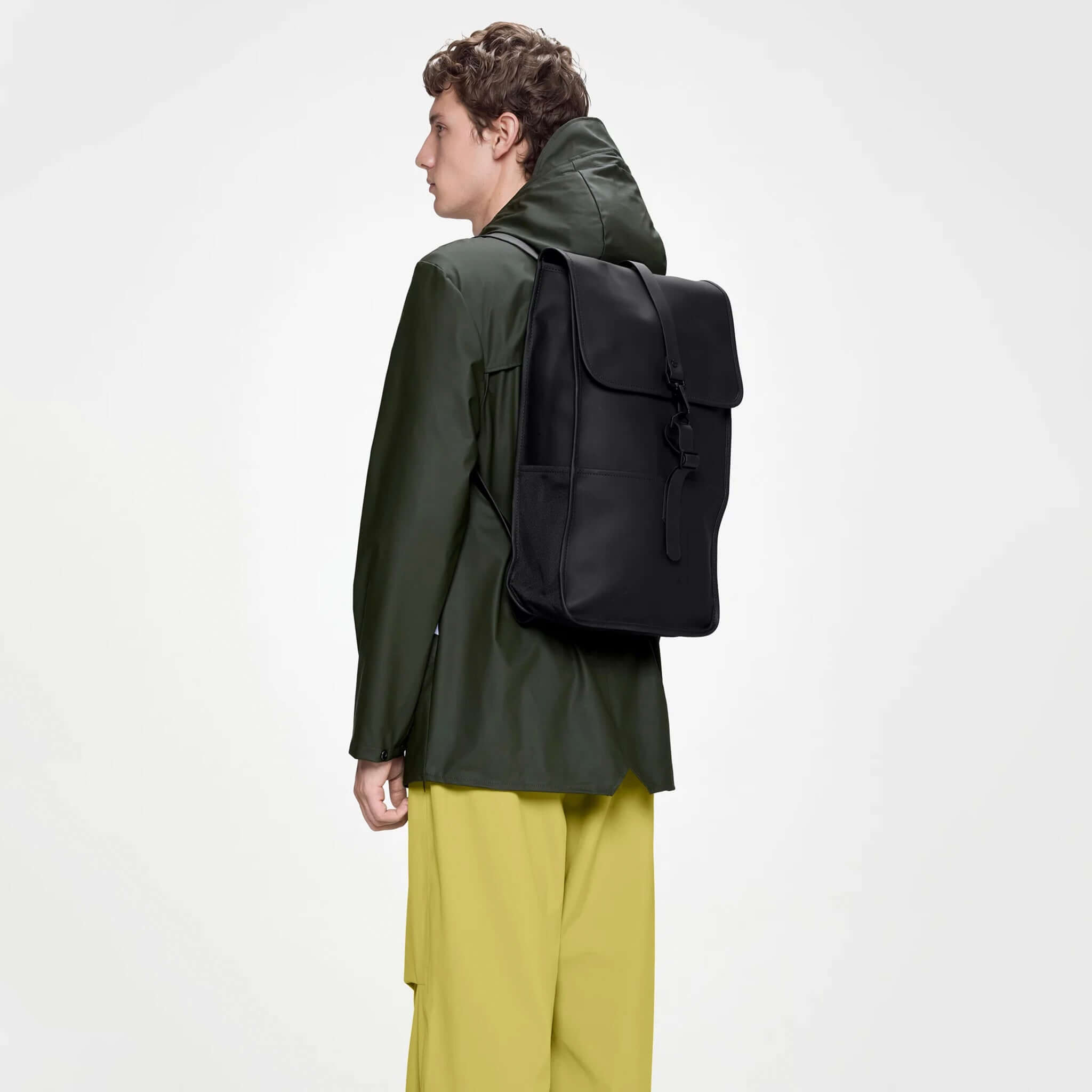 Rains Backpack - Black worn by a model in a green jacket and yellow pants, showcasing a sleek and timeless design.