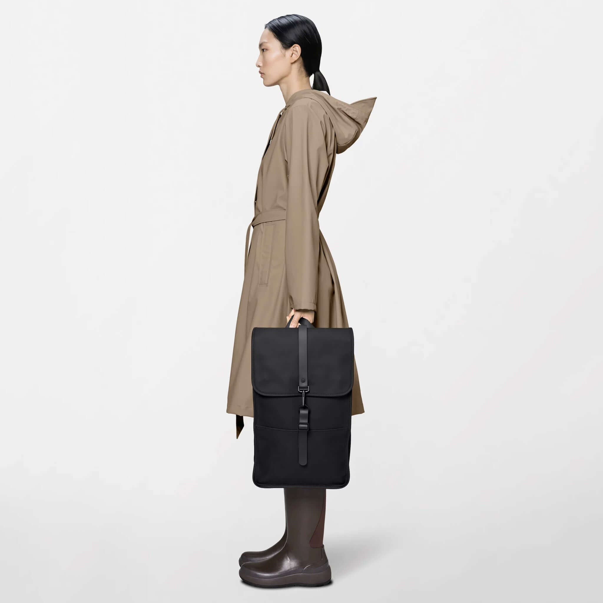 Rains Backpack - Black being held by a model in a brown coat, showcasing its sleek design and functionality.