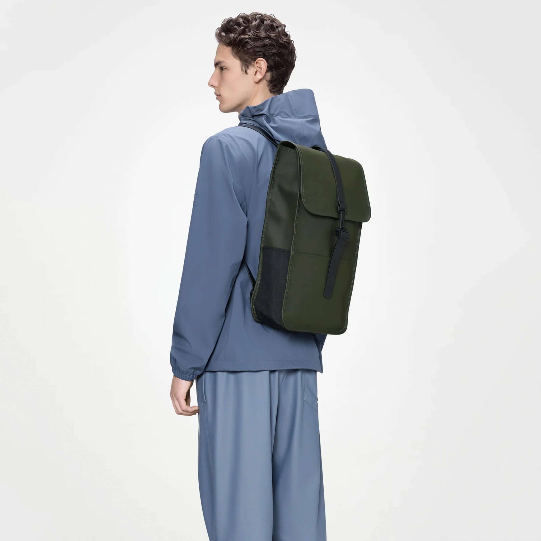 Rains Backpack - Green showcased on a model wearing a blue jacket, highlighting its sleek design and waterproof fabric.