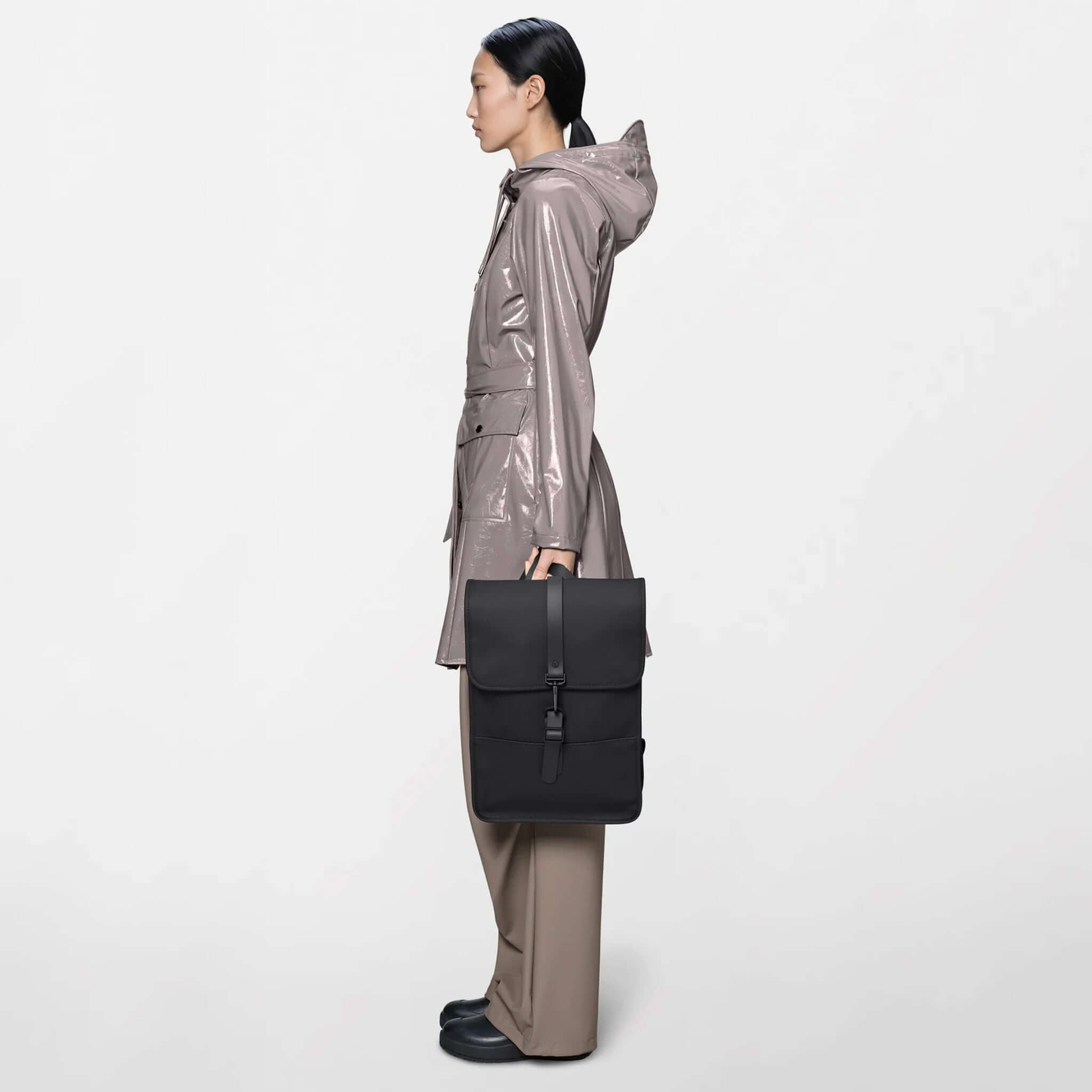 Woman wearing a raincoat and holding the Rains Backpack Mini - Black, showcasing its sleek design.