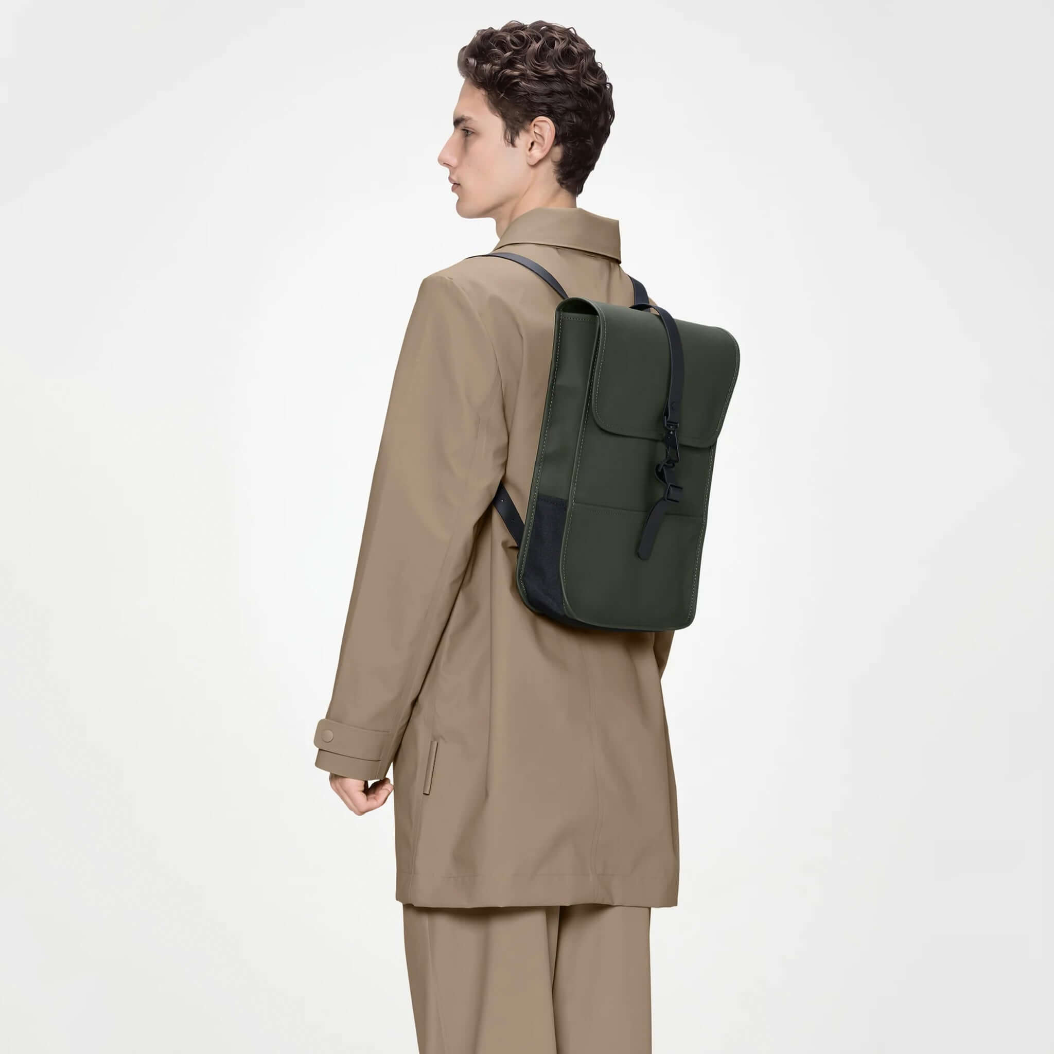 Rains Backpack Mini - Green worn by a model in a beige coat from the back, showcasing its sleek design.