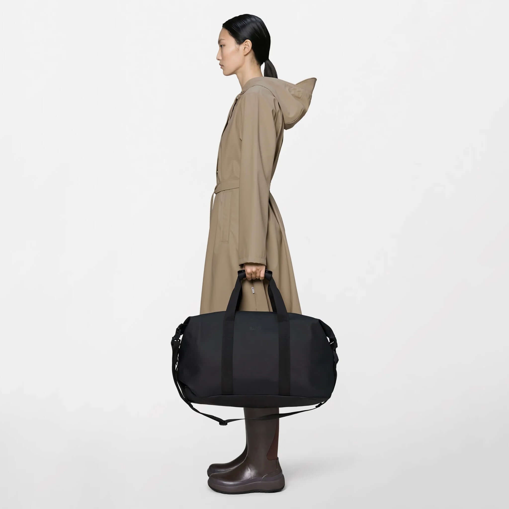 Rains Hilo Weekend Bag - Black held by a woman in a raincoat, showcasing its sleek design.