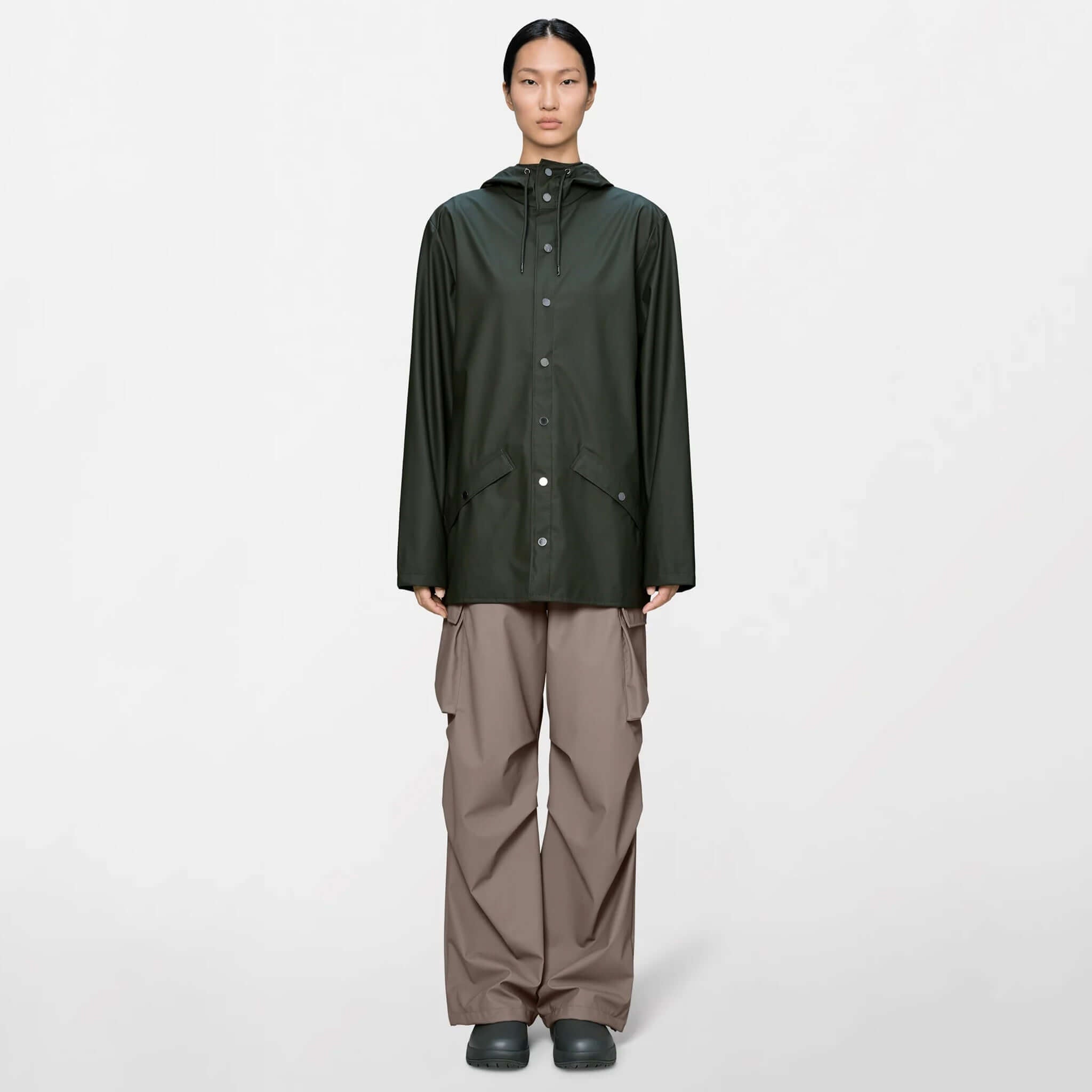 A model wearing the Rains Jacket - Green, a minimalistic rain jacket with adjustable hood and pockets.