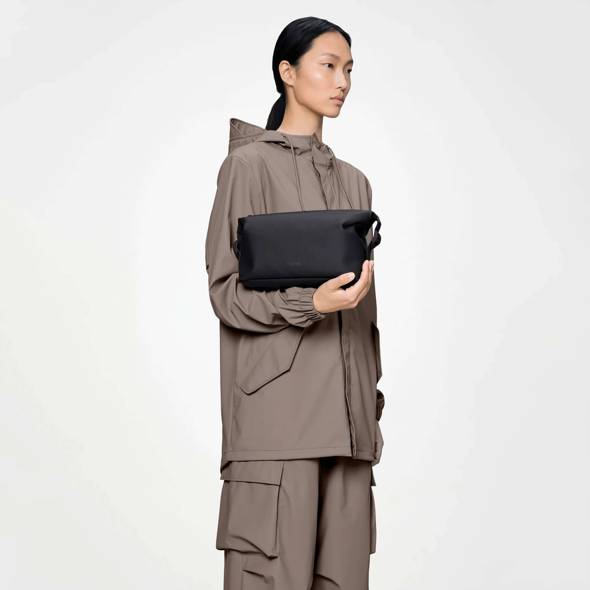 Rains Wash Bag Small - Black held by a model wearing a beige rain jacket and matching pants.