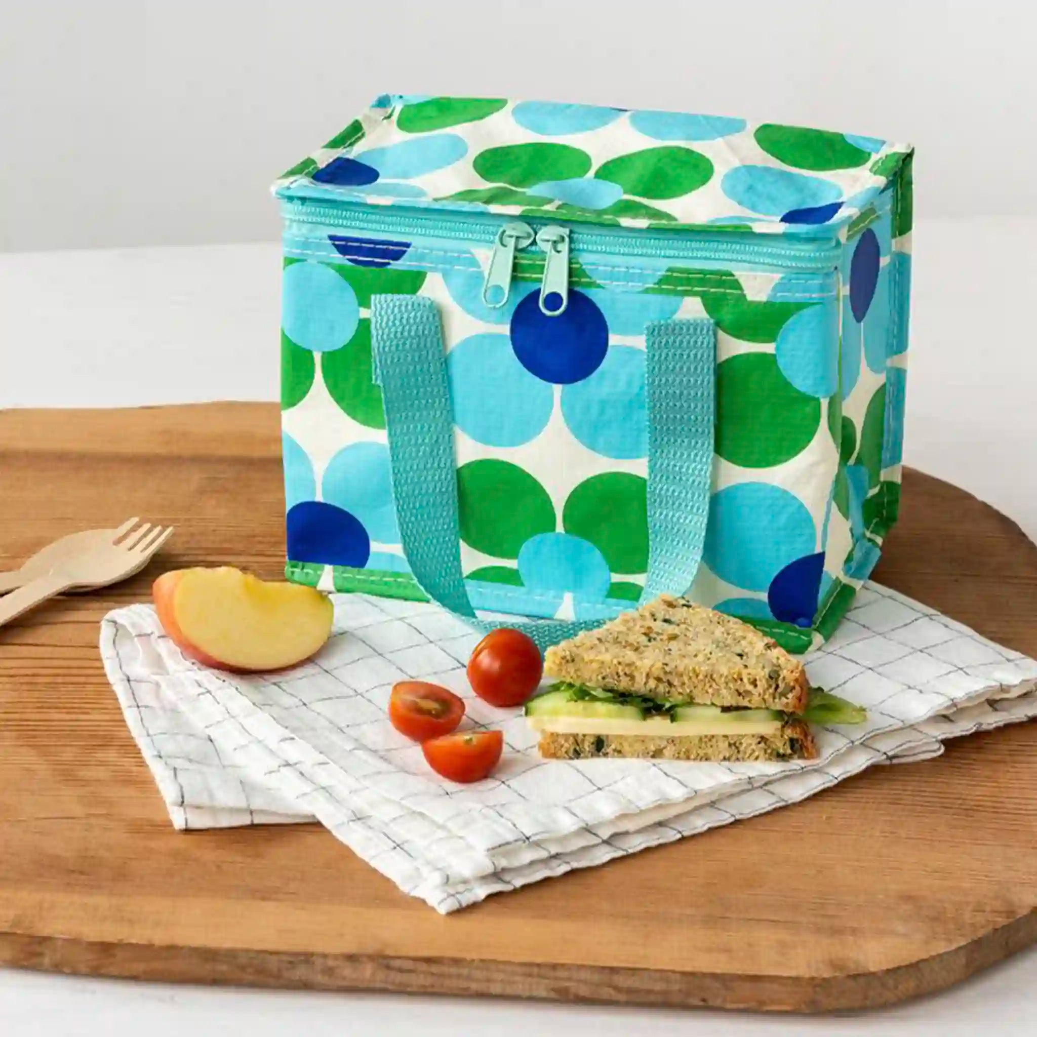 insulated lunch bag with green and blue daisy pattern on a wooden table with a sandwich and fruit.