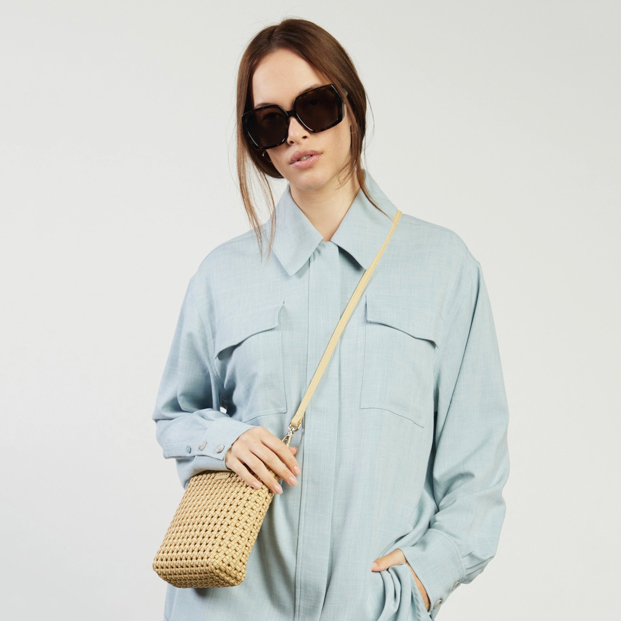 Woman wearing a light blue shirt and sunglasses, holding a woven handbag against a white background