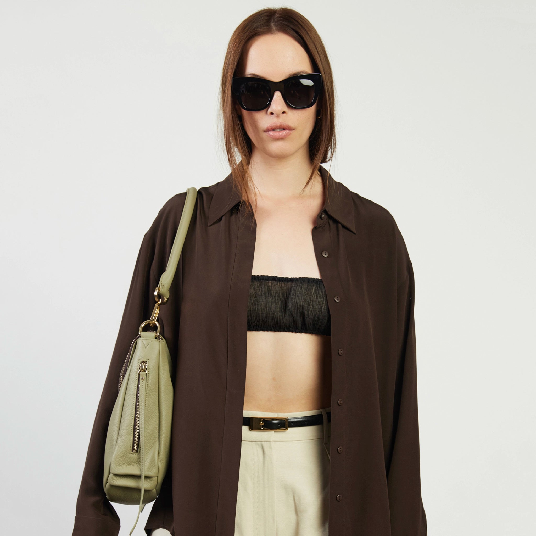 Woman wearing sunglasses, a brown shirt, black bralette, and white pants with a green bag on a plain background