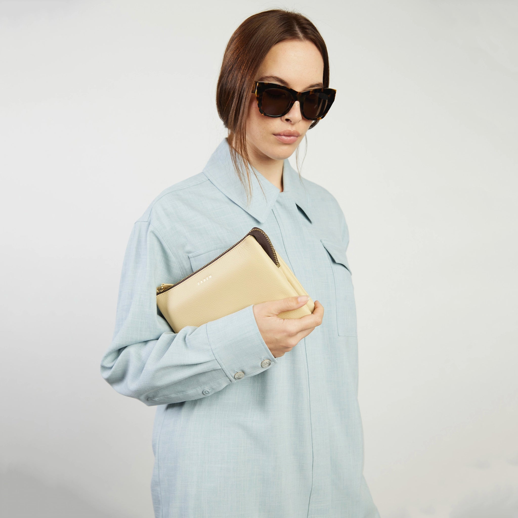 Woman wearing a light blue coat and sunglasses, holding a beige clutch against a plain background
