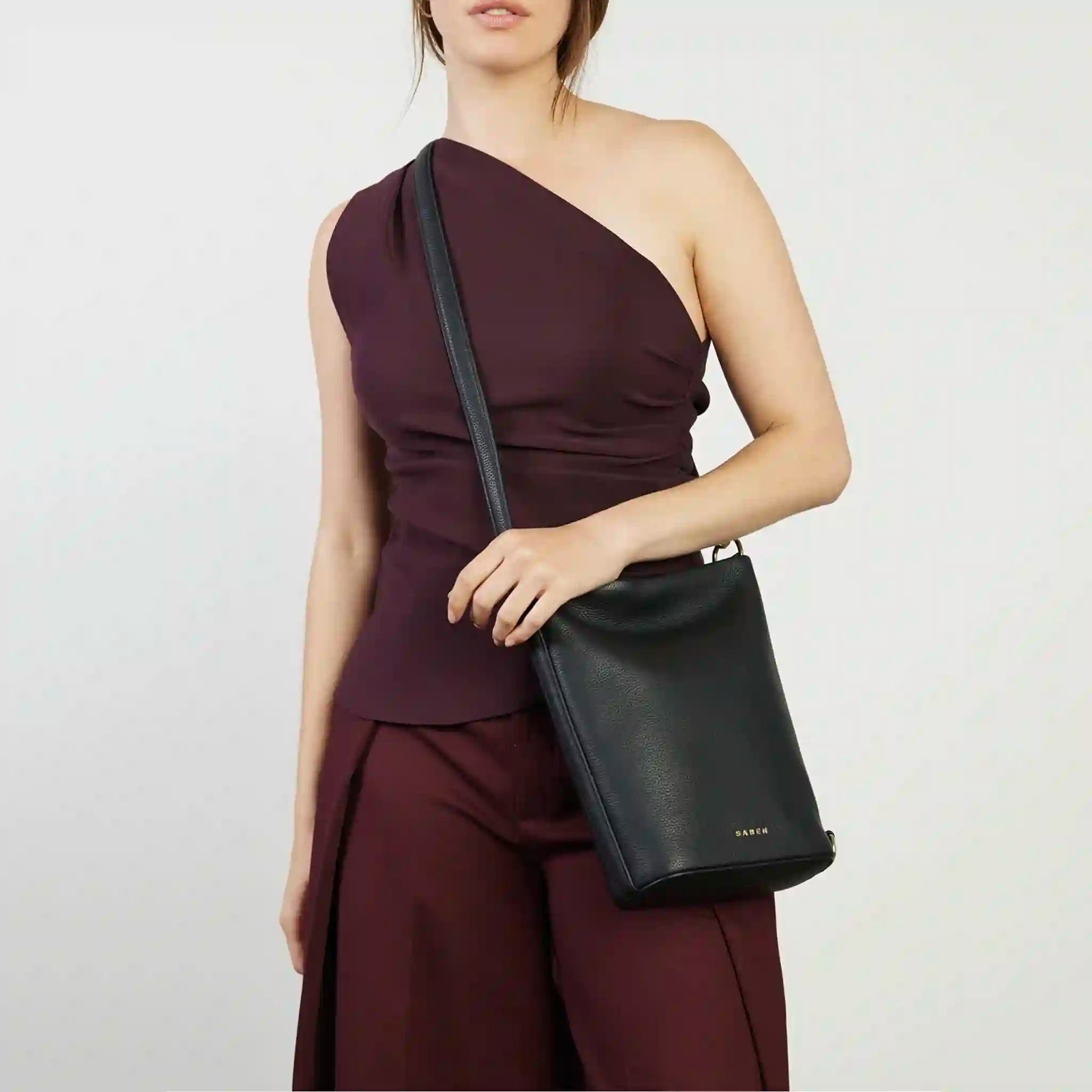 Woman wearing a burgundy one-shoulder dress holding a black handbag against a white background