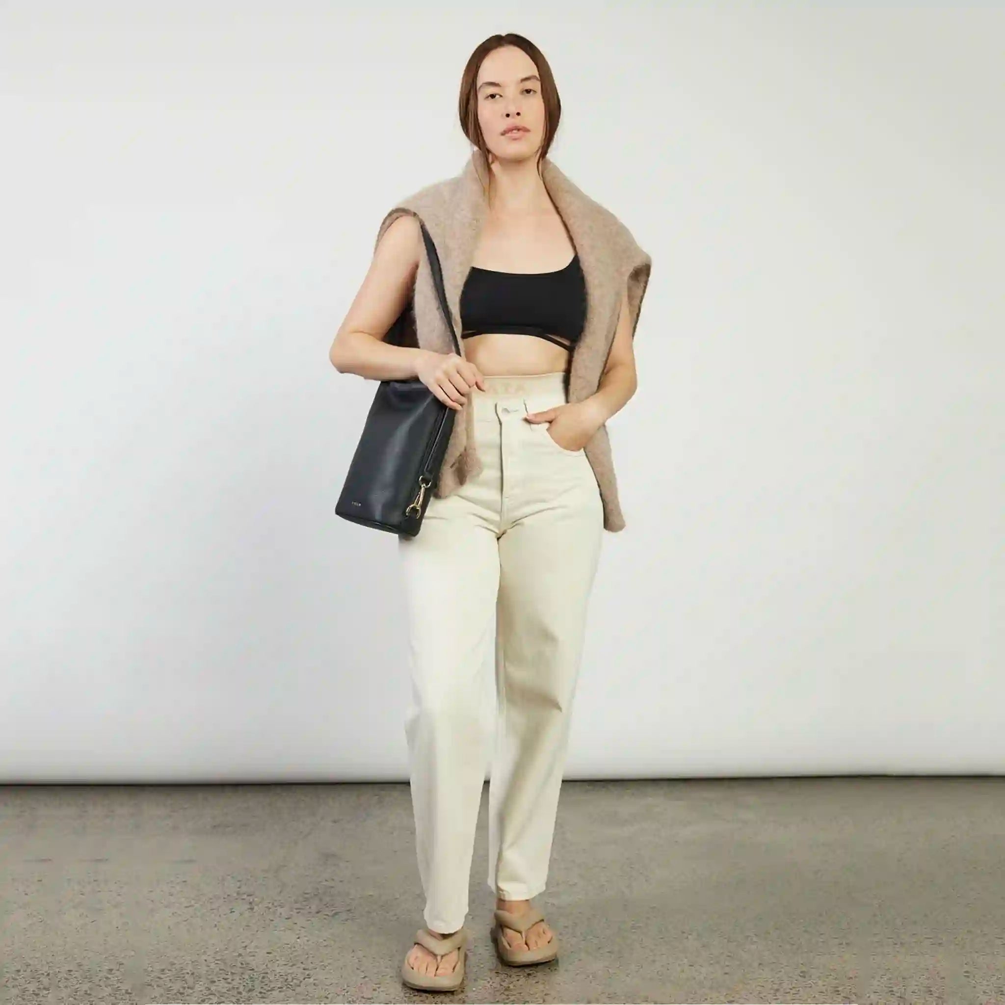 Woman wearing a black top, beige vest, white pants, and brown shoes against a plain background