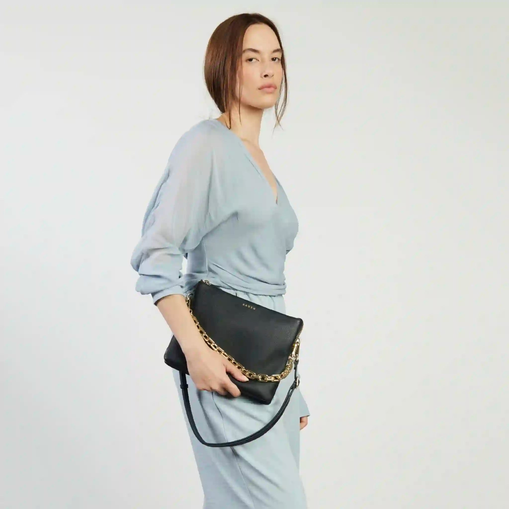 Woman holding a black handbag with gold chain detail against a plain background