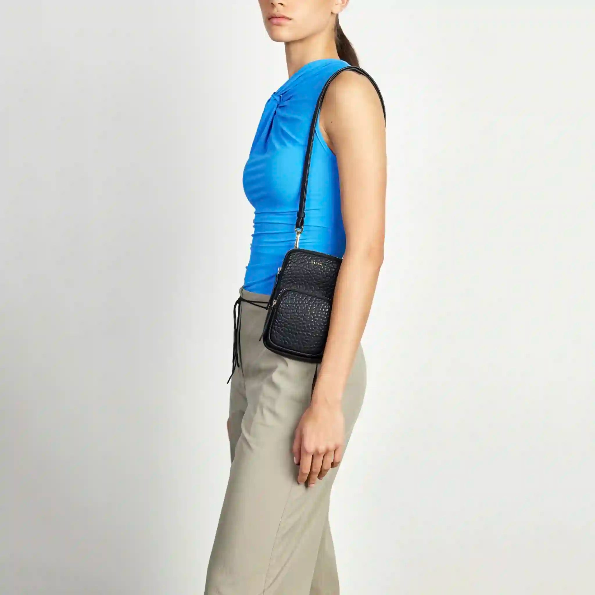 Person wearing a blue top and beige pants with a black crossbody bag on a white background