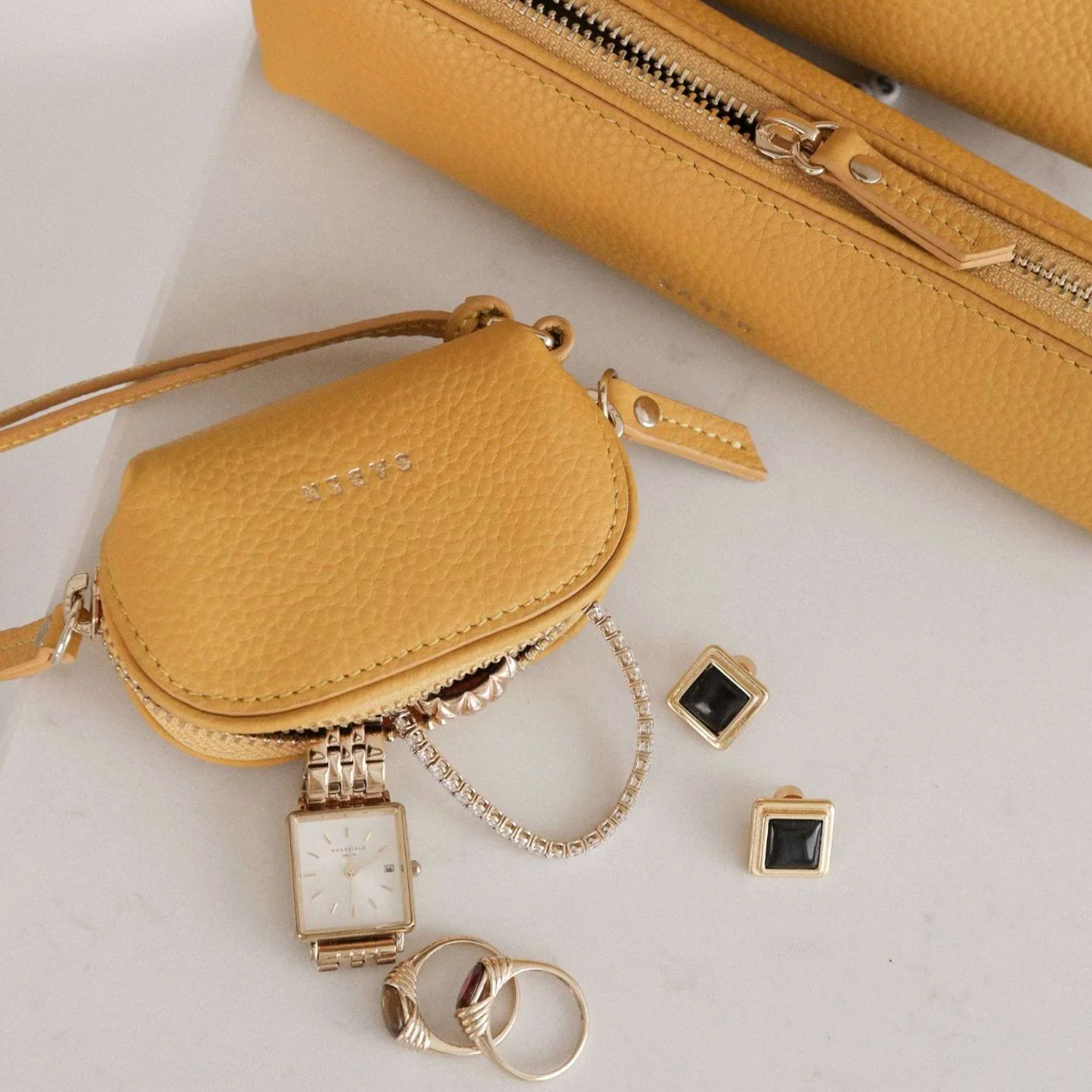 Yellow handbag with accessories including a watch, rings, and earrings on a light background