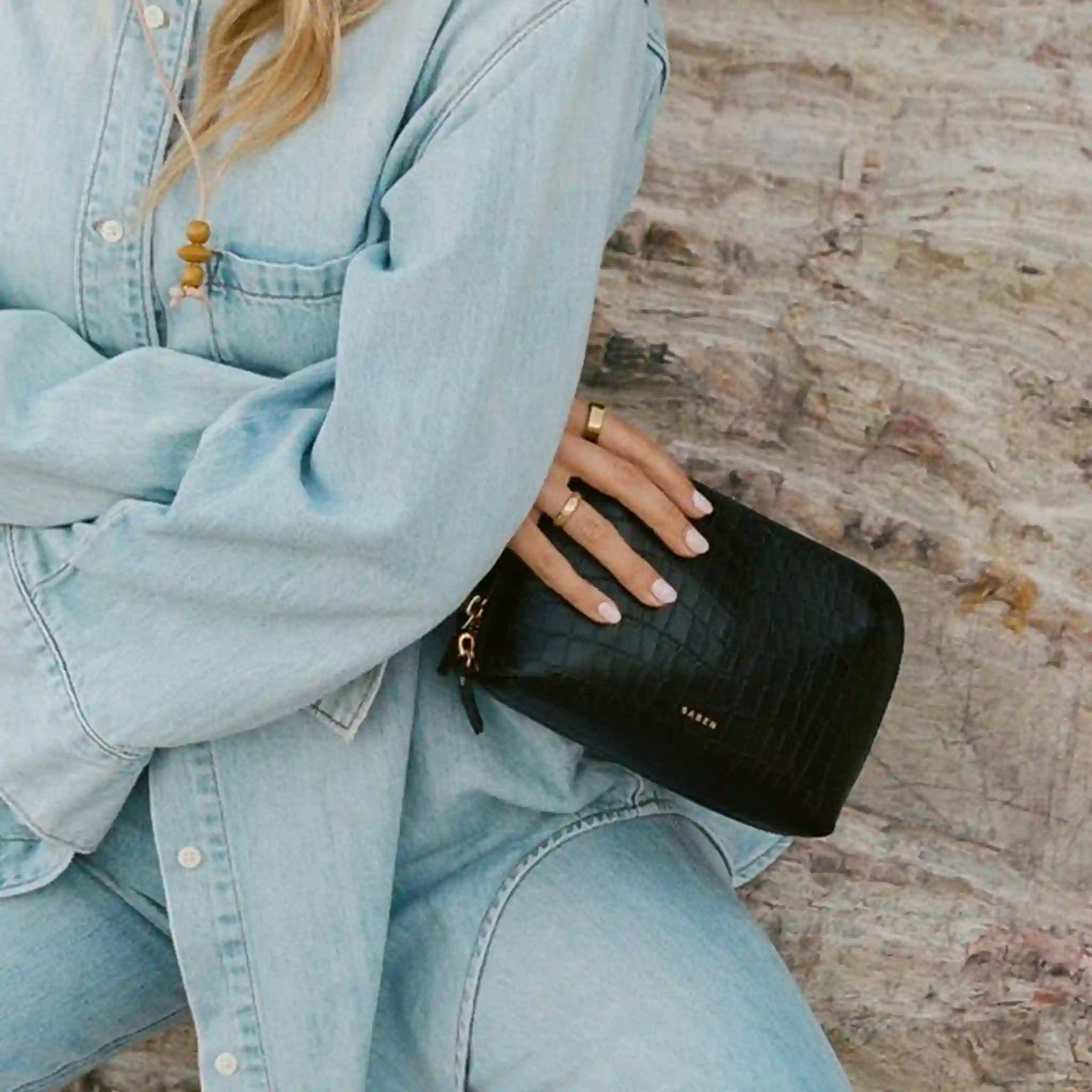 Person wearing a light blue denim jacket holding a black clutch with 'Khaite' branding.
