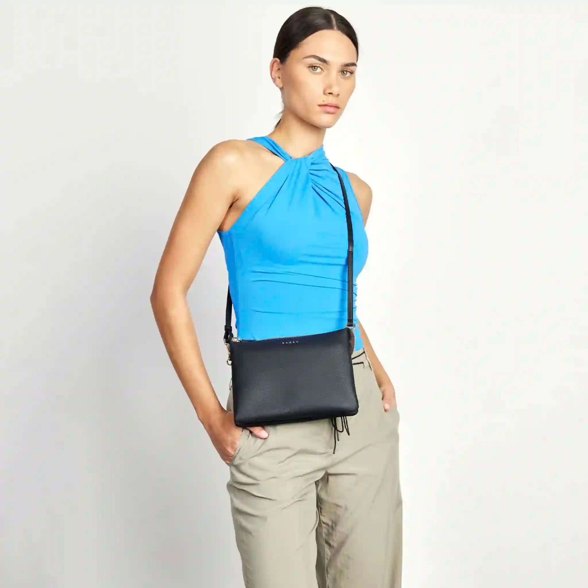 Woman wearing a blue top and beige pants with a black crossbody bag on a light gray background