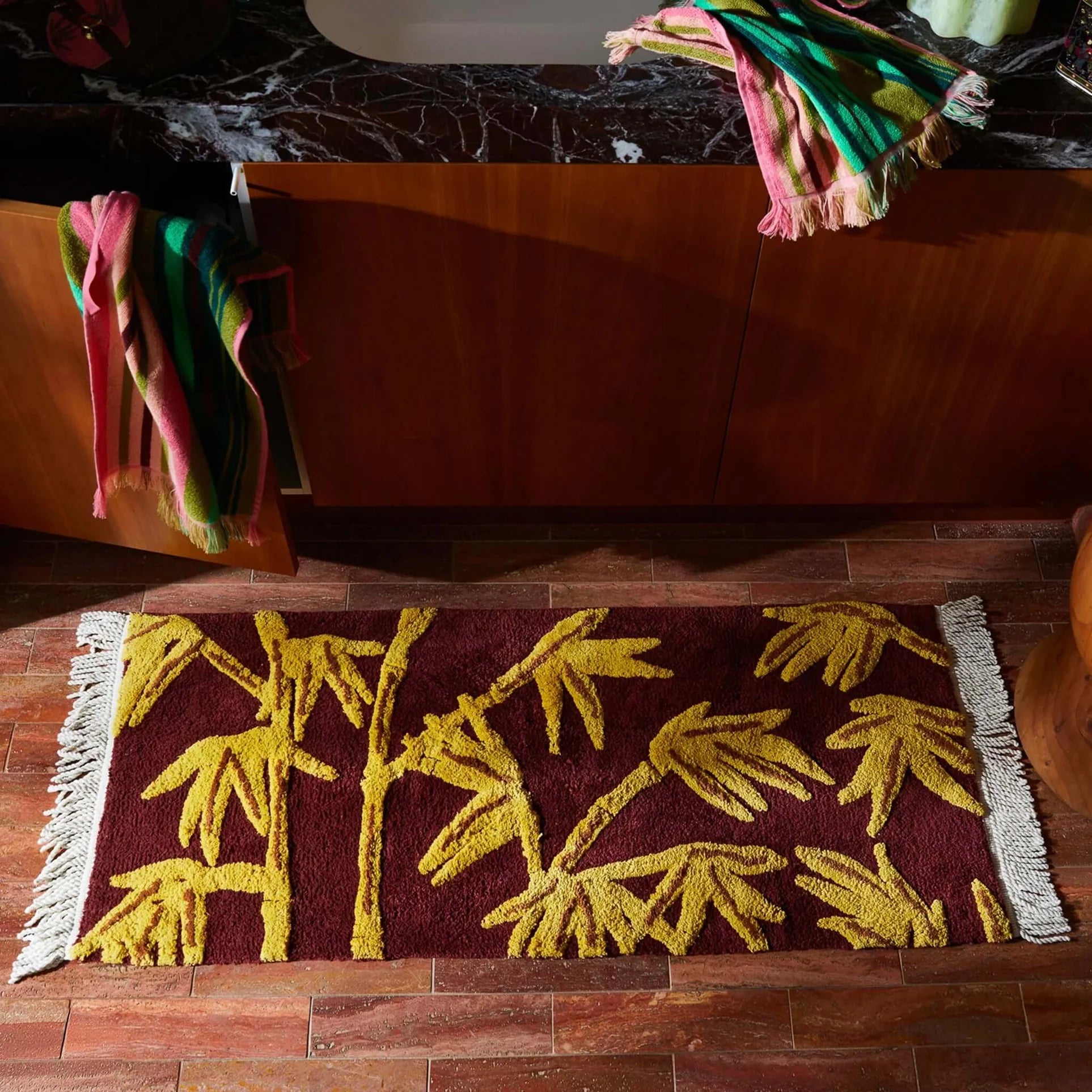 A rectangular bath mat with fringe ends lies on a wood floor in front of a wooden cabinet. The mat has a deep burgundy background with raised yellow bamboo motifs.
