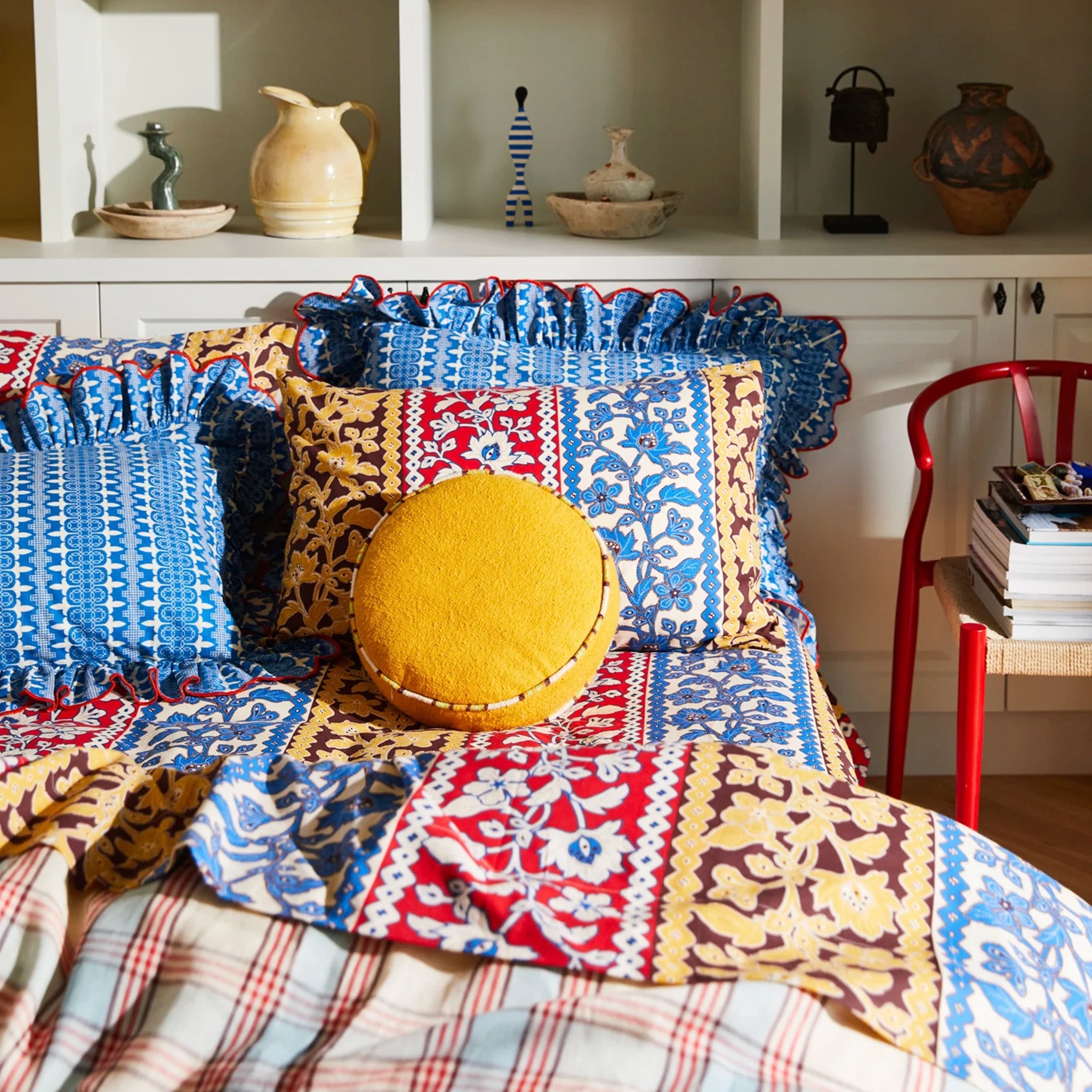 Colorful patterned cushions on a bed with a shelf of decorative items in the background.