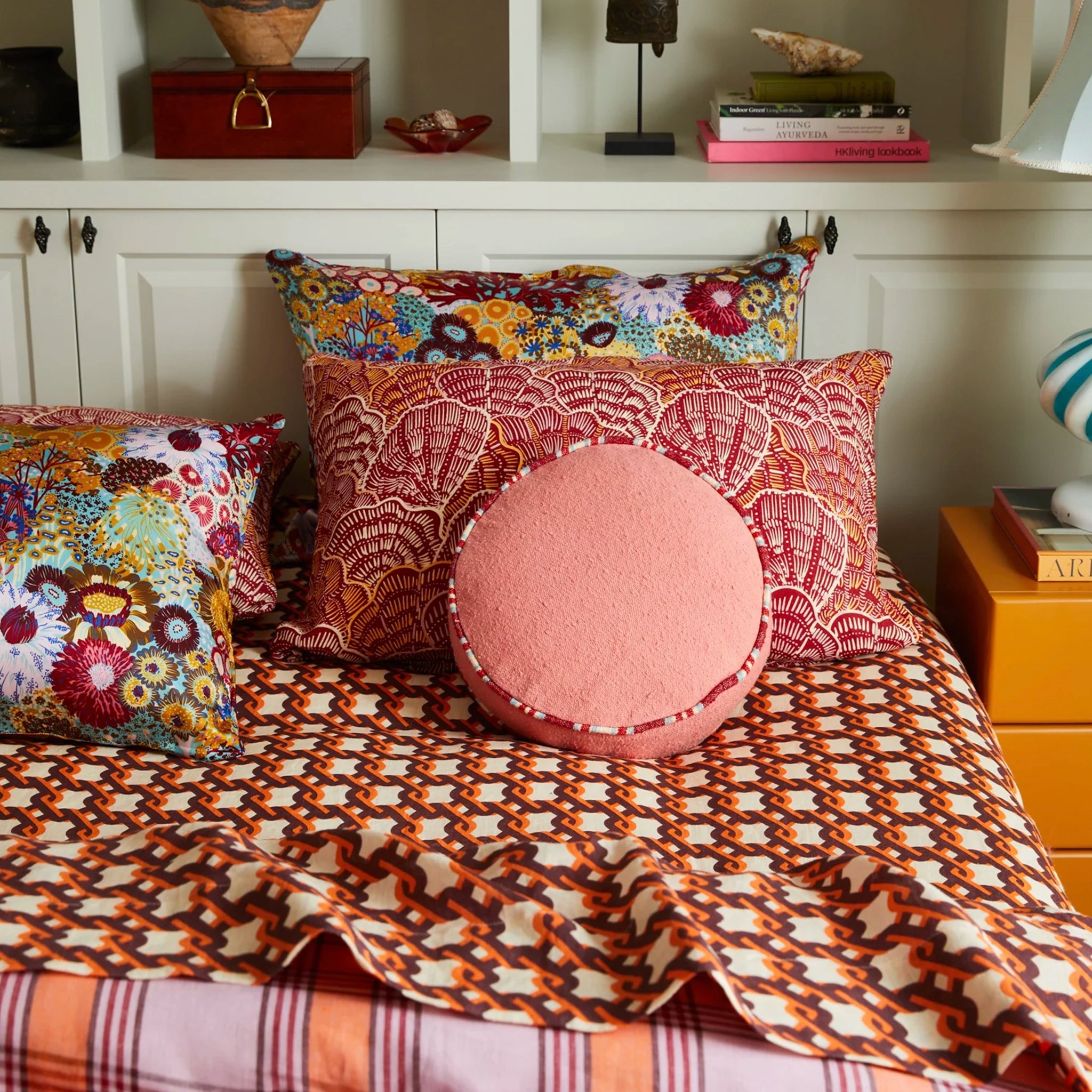 Patterned pillows and a round pink cushion on a bed with a shelf in the background.
