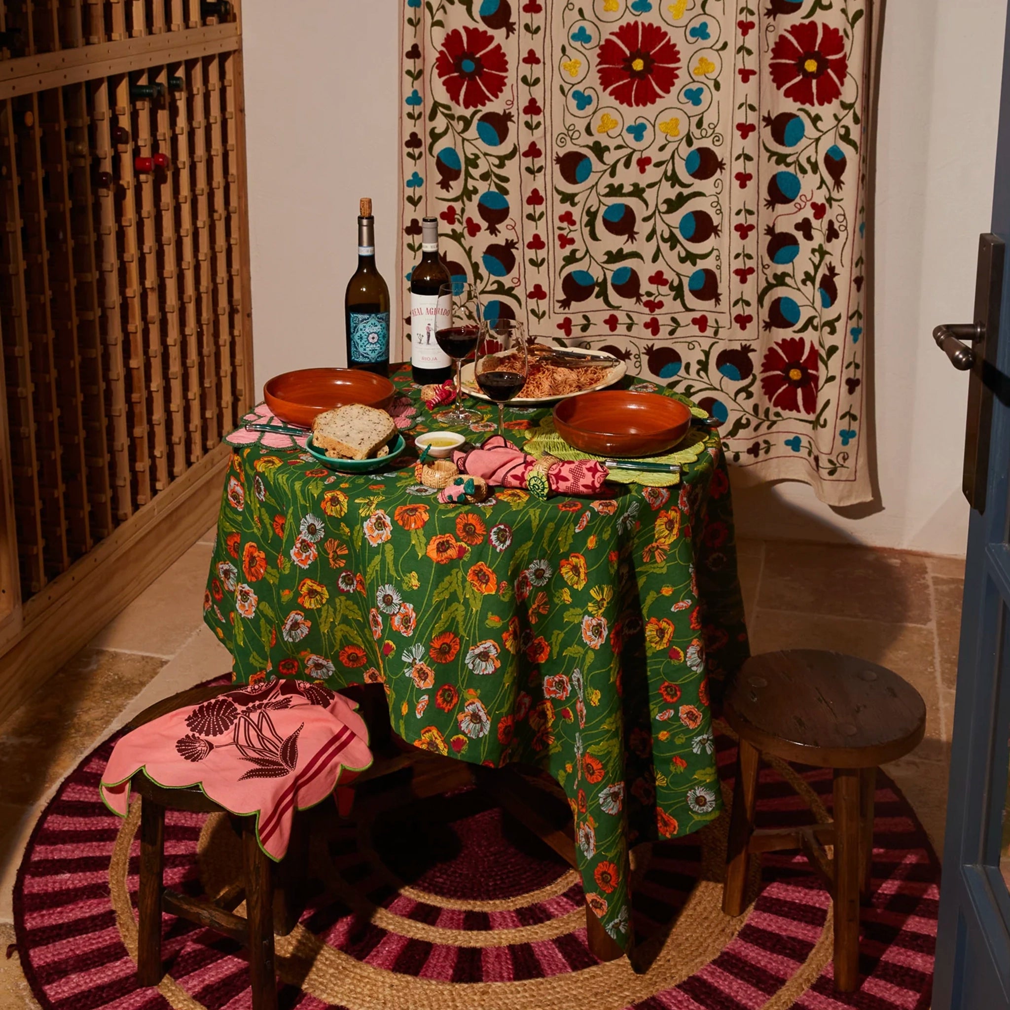 Dining table set with food and wine bottles, floral tablecloth, and decorative chairs in a room with a patterned wall hanging.