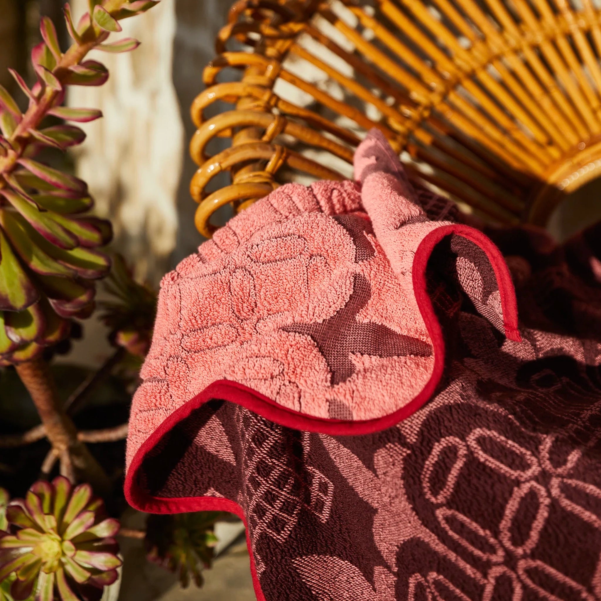 Pink textured towel with red border draped over a wicker chair with plants in the background
