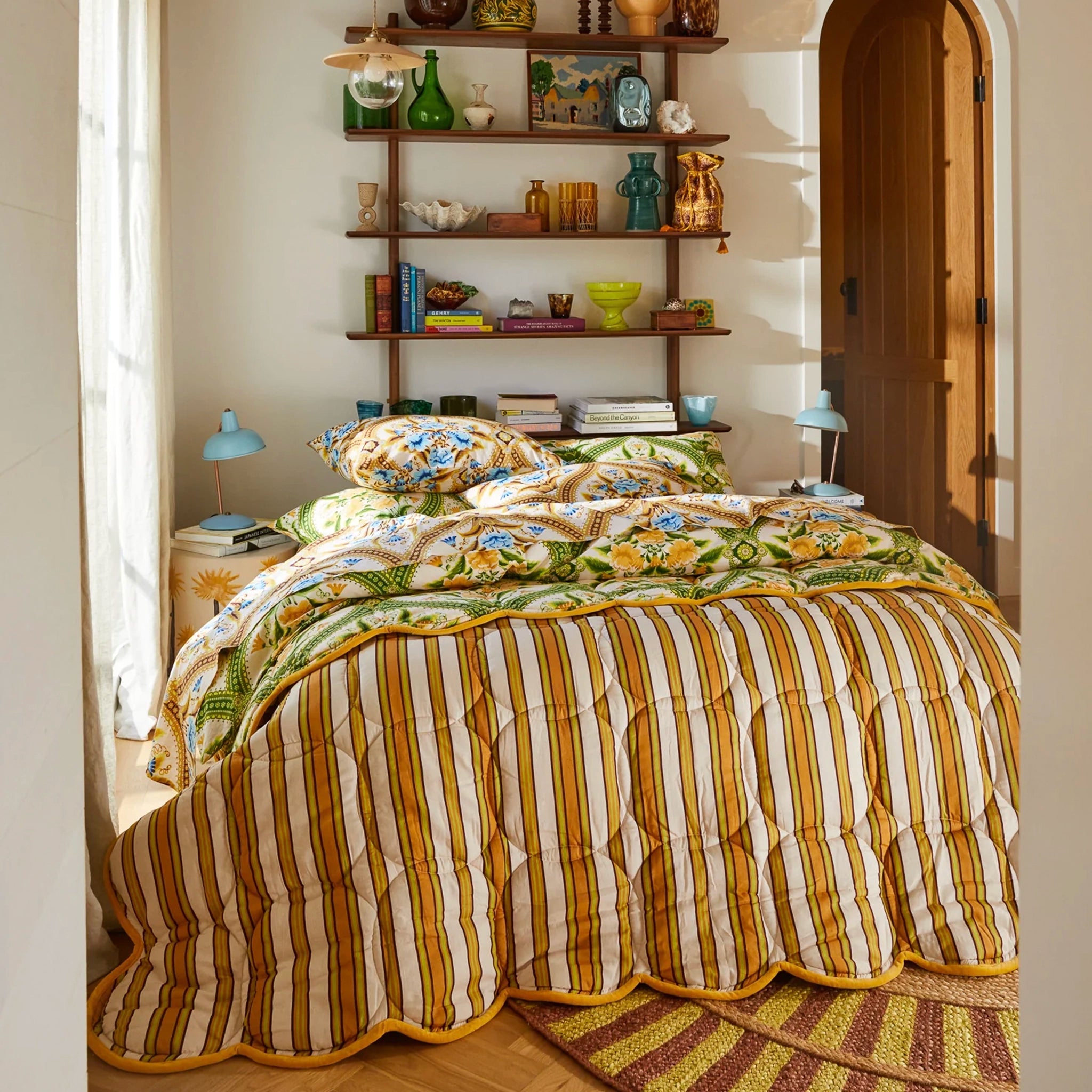 Bedroom with striped bedding and decorative pillows, featuring a bookshelf and lamp.