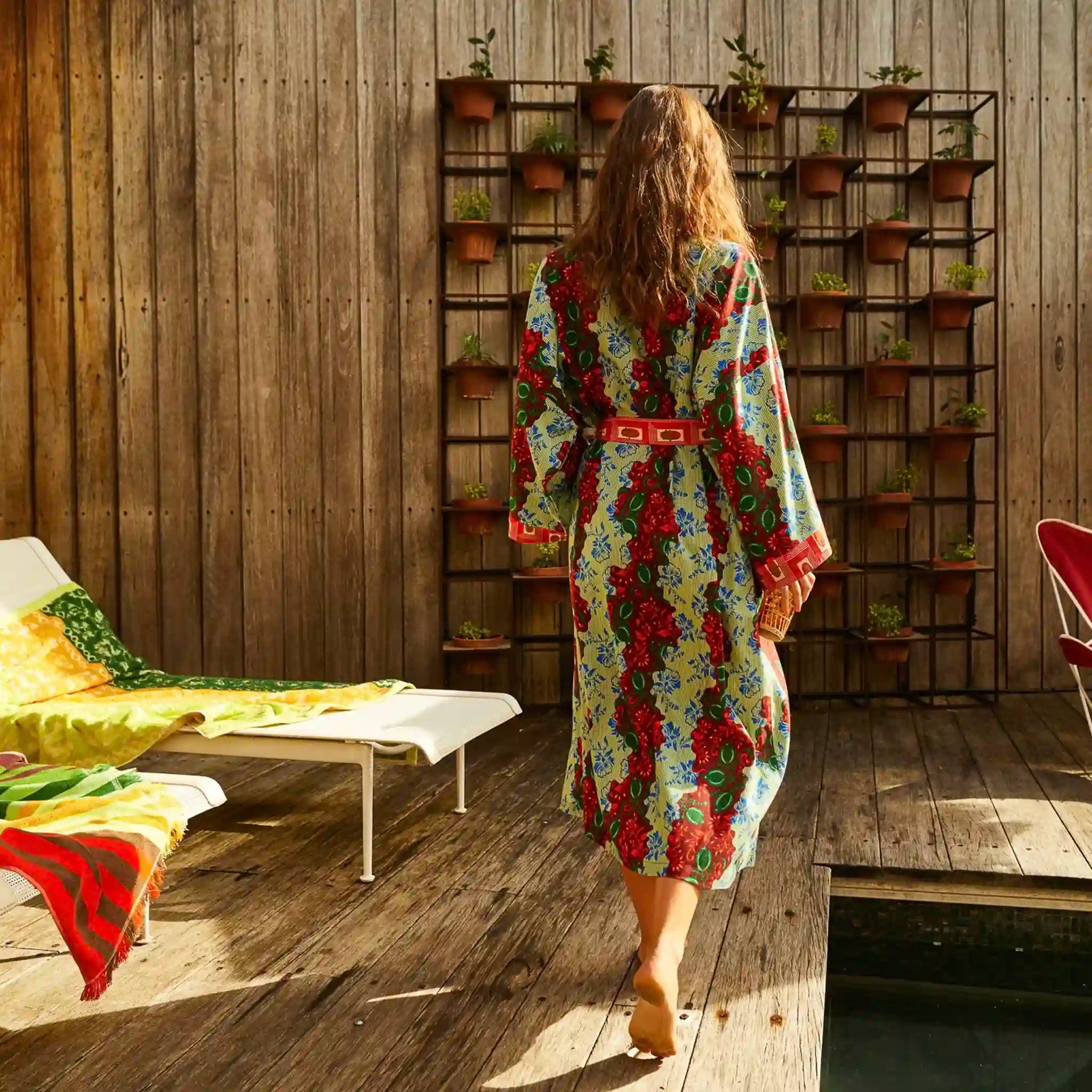 Woman in a colorful floral robe walking towards a wooden wall with planters.