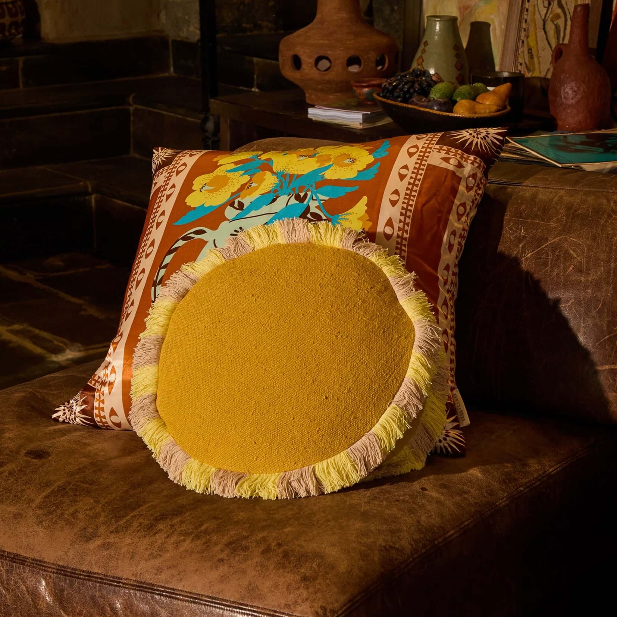 Sage x Clare Reese Round Cushion - Ochre with lemon and blush fringe on a brown sofa.